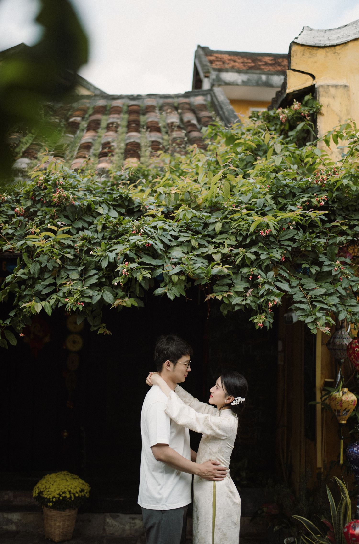 A couple stands close together under lush green foliage in a traditional Asian garden setting.