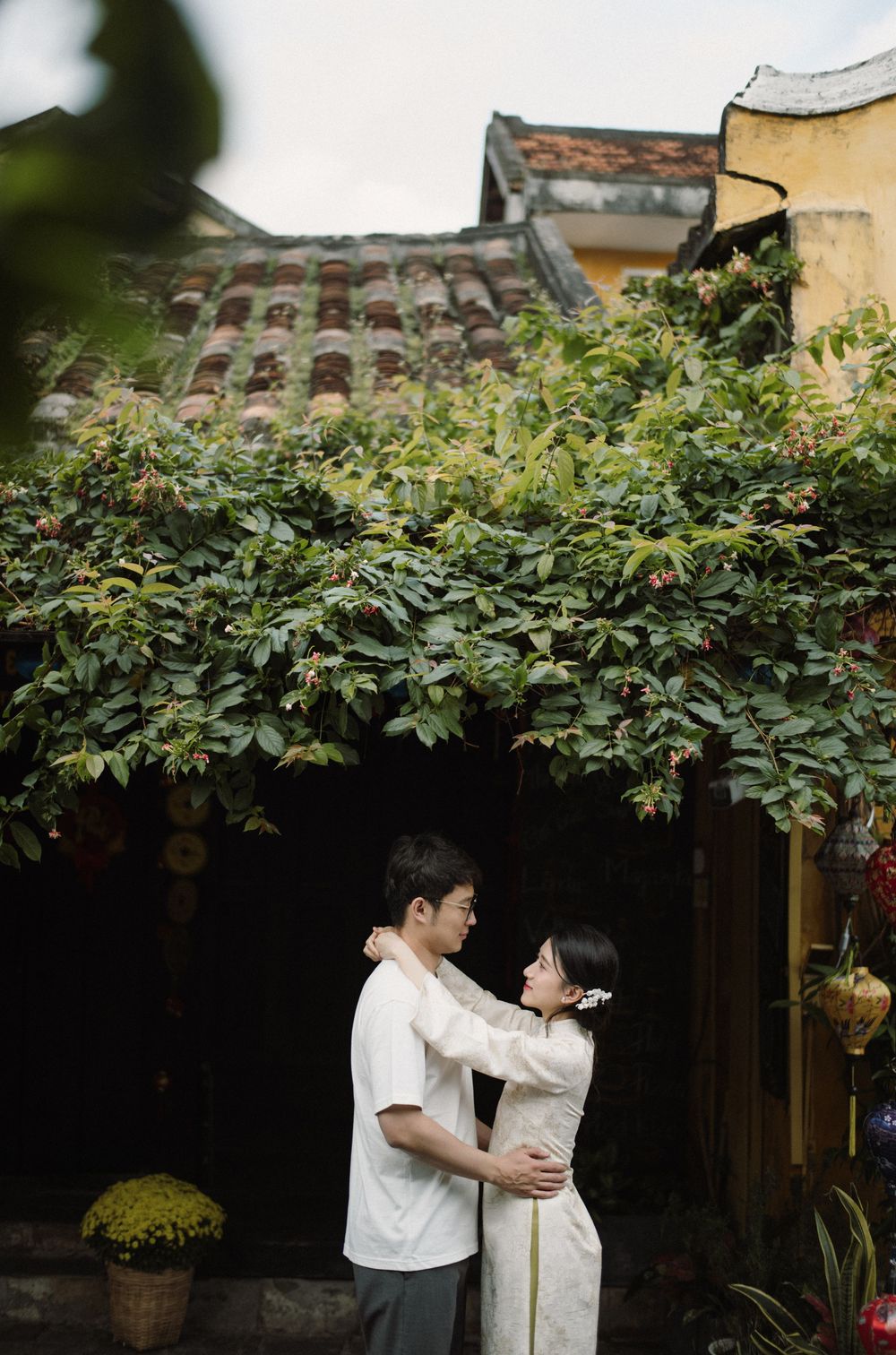 A couple stands close together under lush green foliage in a traditional Asian garden setting.