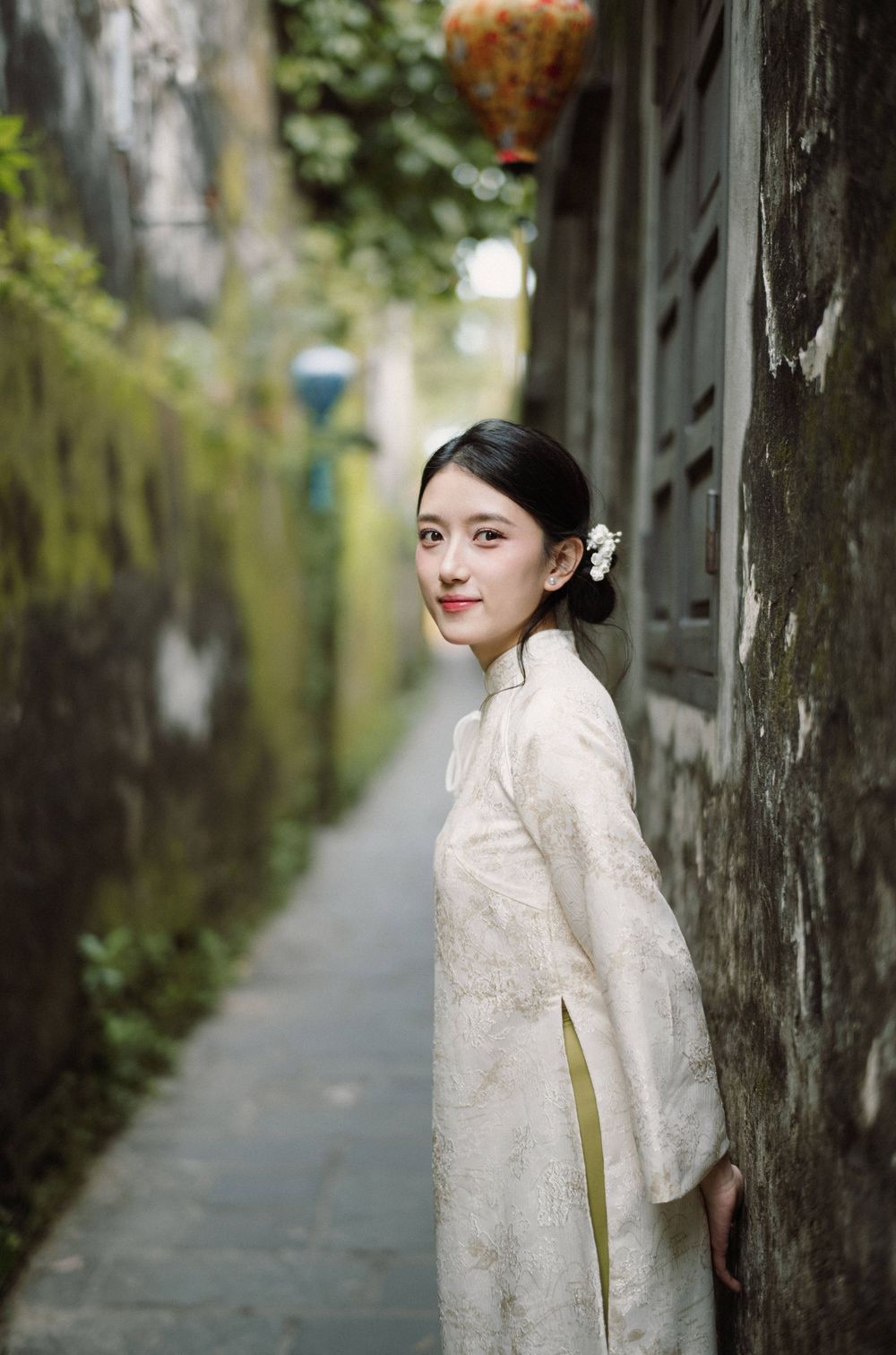 Figure in white lace dress poses in an atmospheric alley with moss-covered walls.