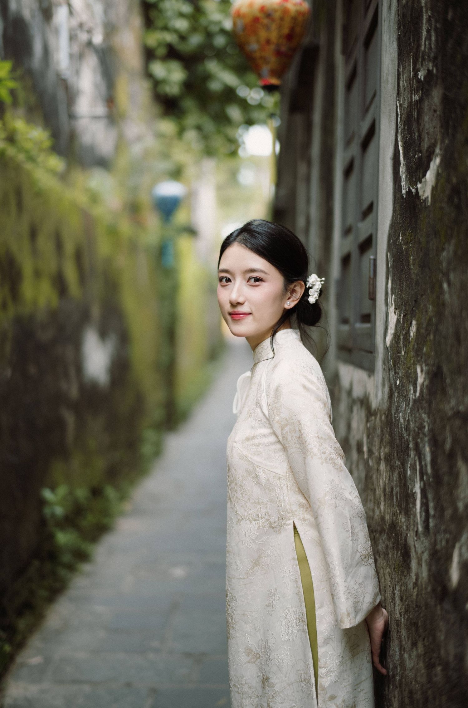 Figure in white lace dress poses in an atmospheric alley with moss-covered walls.