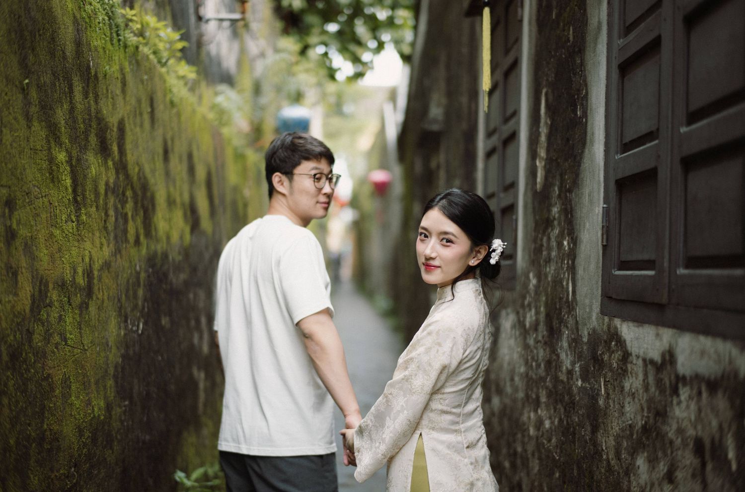 Two people stroll through a historic alley with weathered walls and green moss.