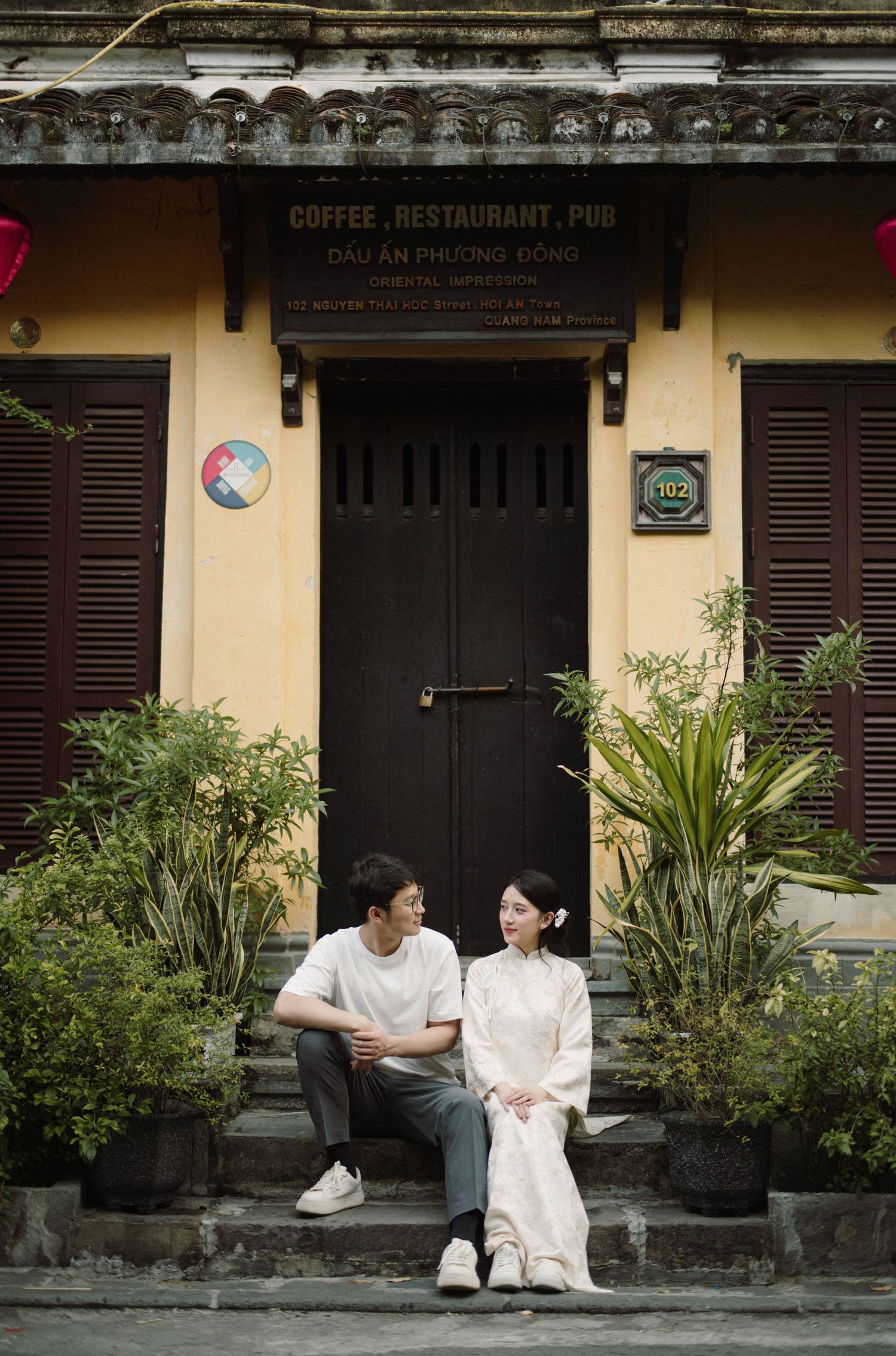 Two people sitting on a bench outside a traditional coffee shop in Vietnam with yellow walls and black shutters.