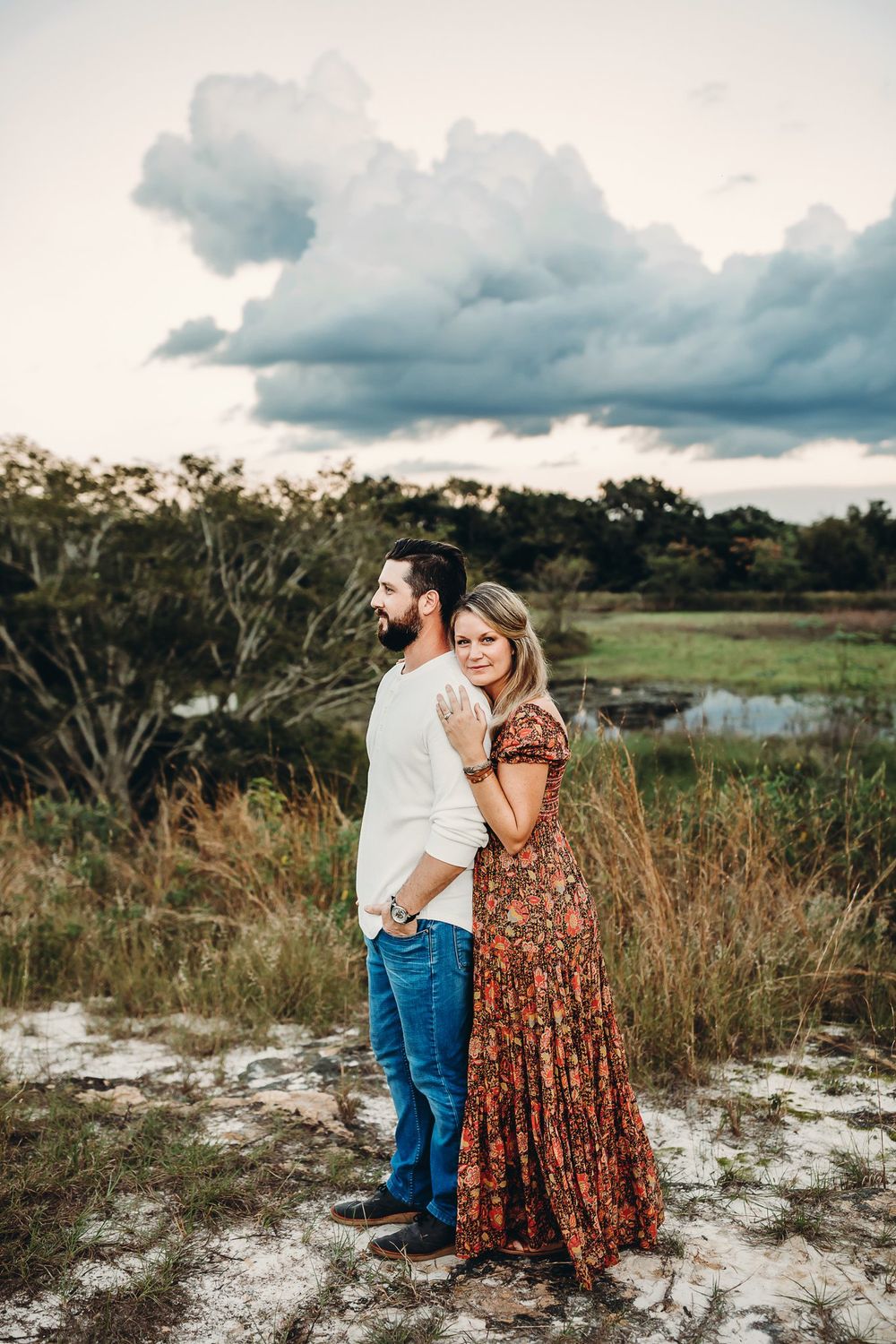 A couple embraces in a romantic photoshoot set against a scenic coastal marsh landscape at sunset.