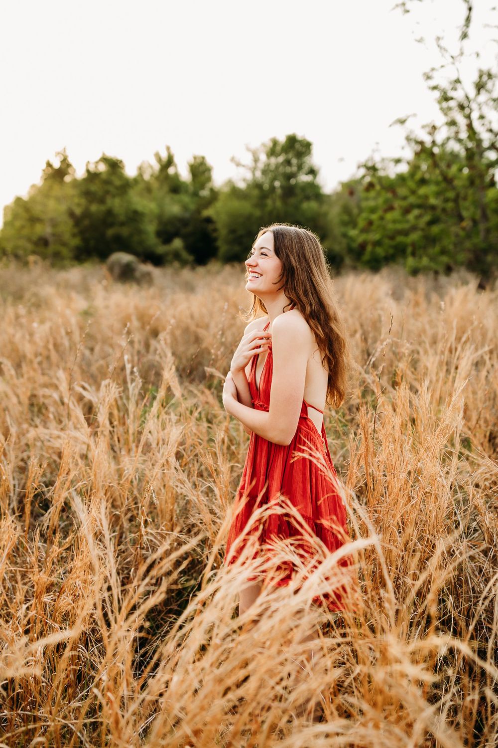 A woman in a flowing red dress stands in a sunlit field of tall golden grass at sunset.