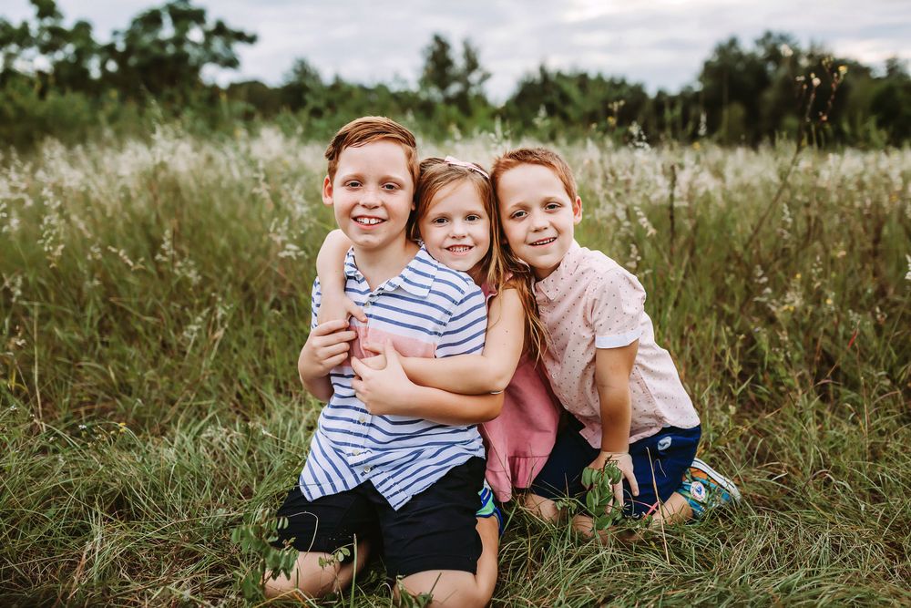 Three children sit close together smiling in a meadow of tall grass on a cloudy day.