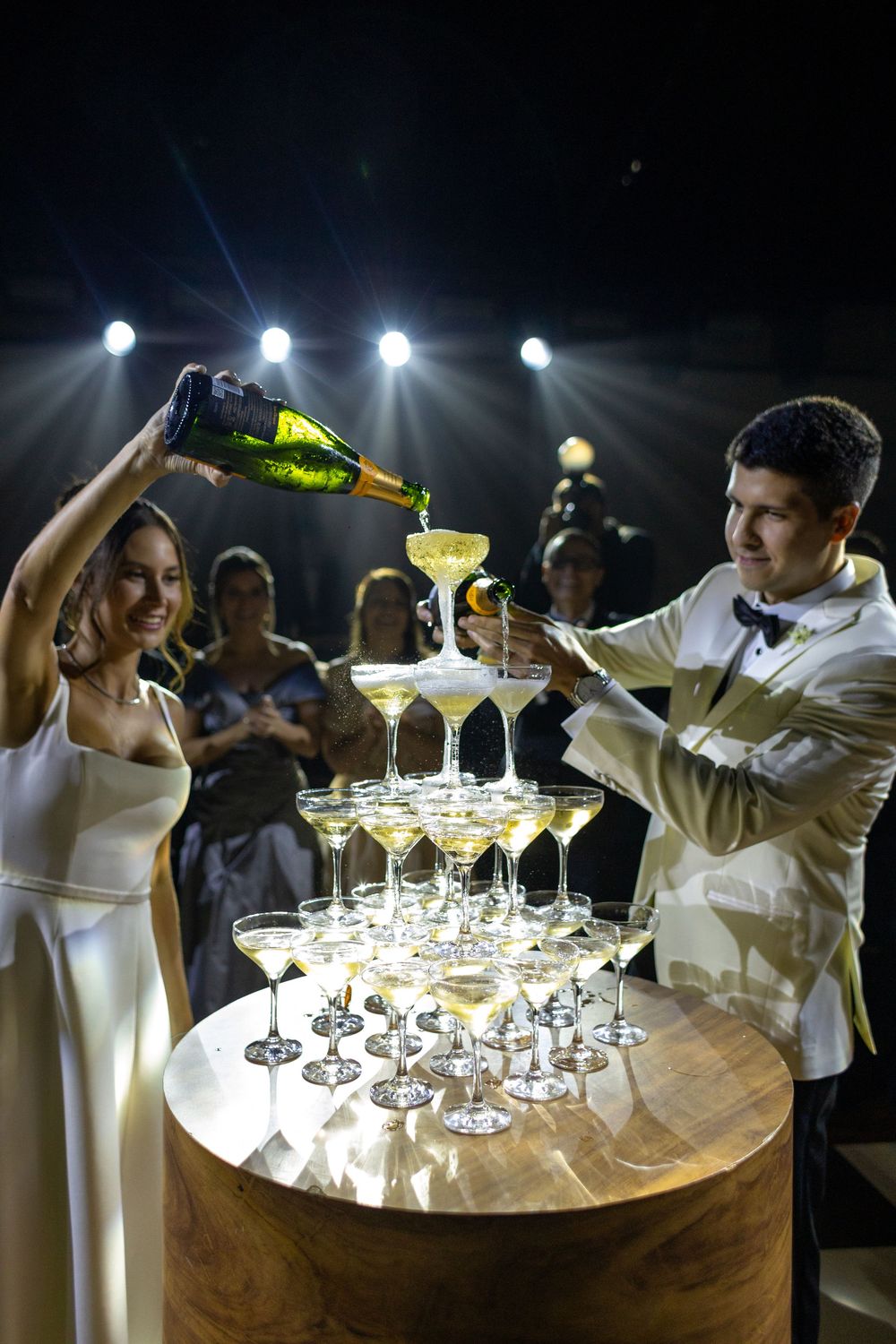 Bartenders in white jackets carefully pour champagne into a tower of crystal glasses under dramatic lighting.