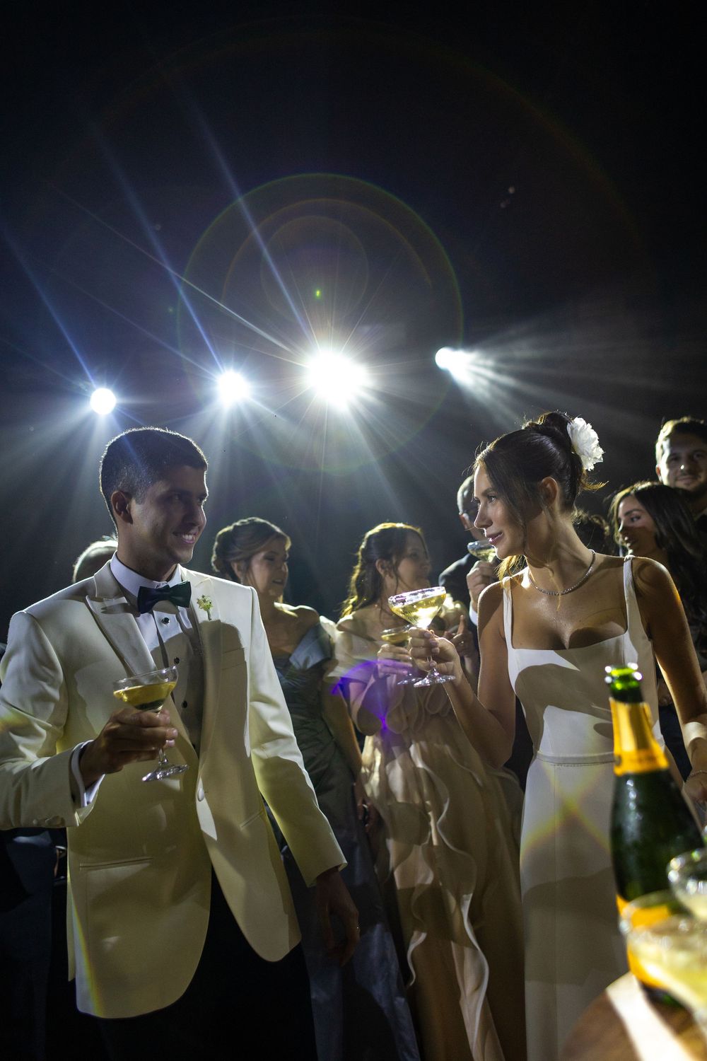 People in formal attire gathered at a nighttime event drinking and socializing under bright lights.