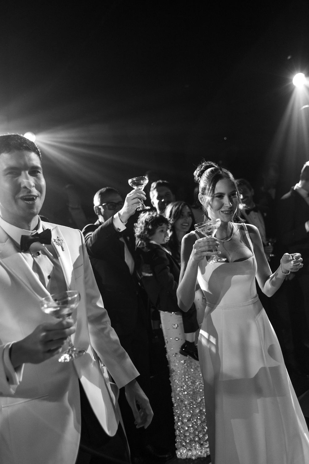 Black and white photo of elegantly dressed wedding guests enjoying cocktails under dramatic lighting.