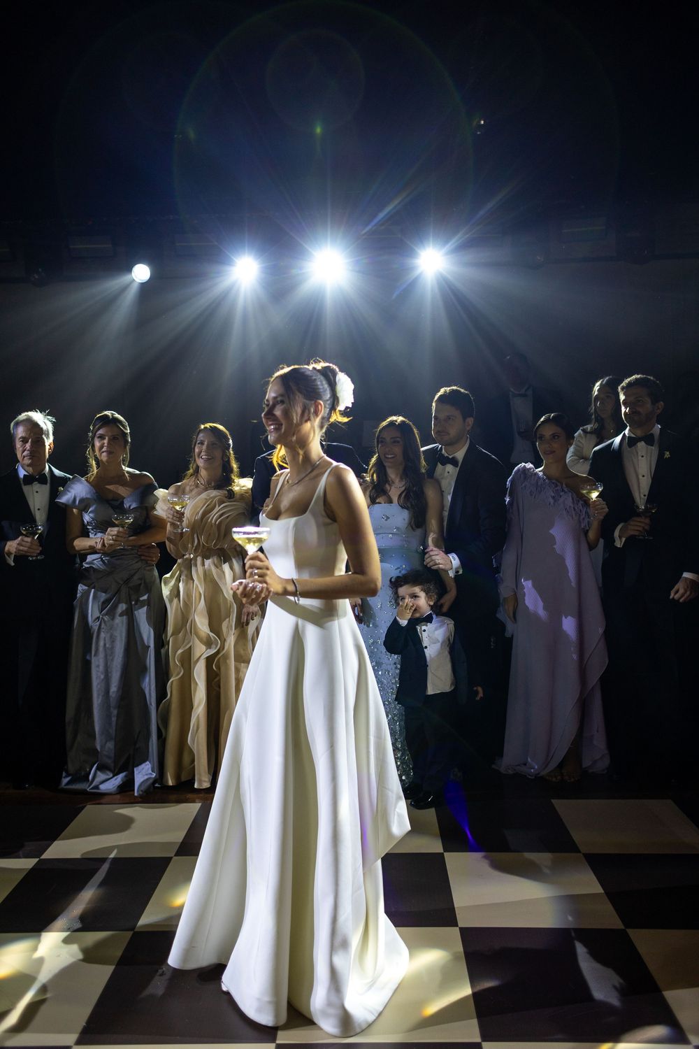 Bride in elegant white gown stands on checkered dance floor under dramatic stage lighting at reception.