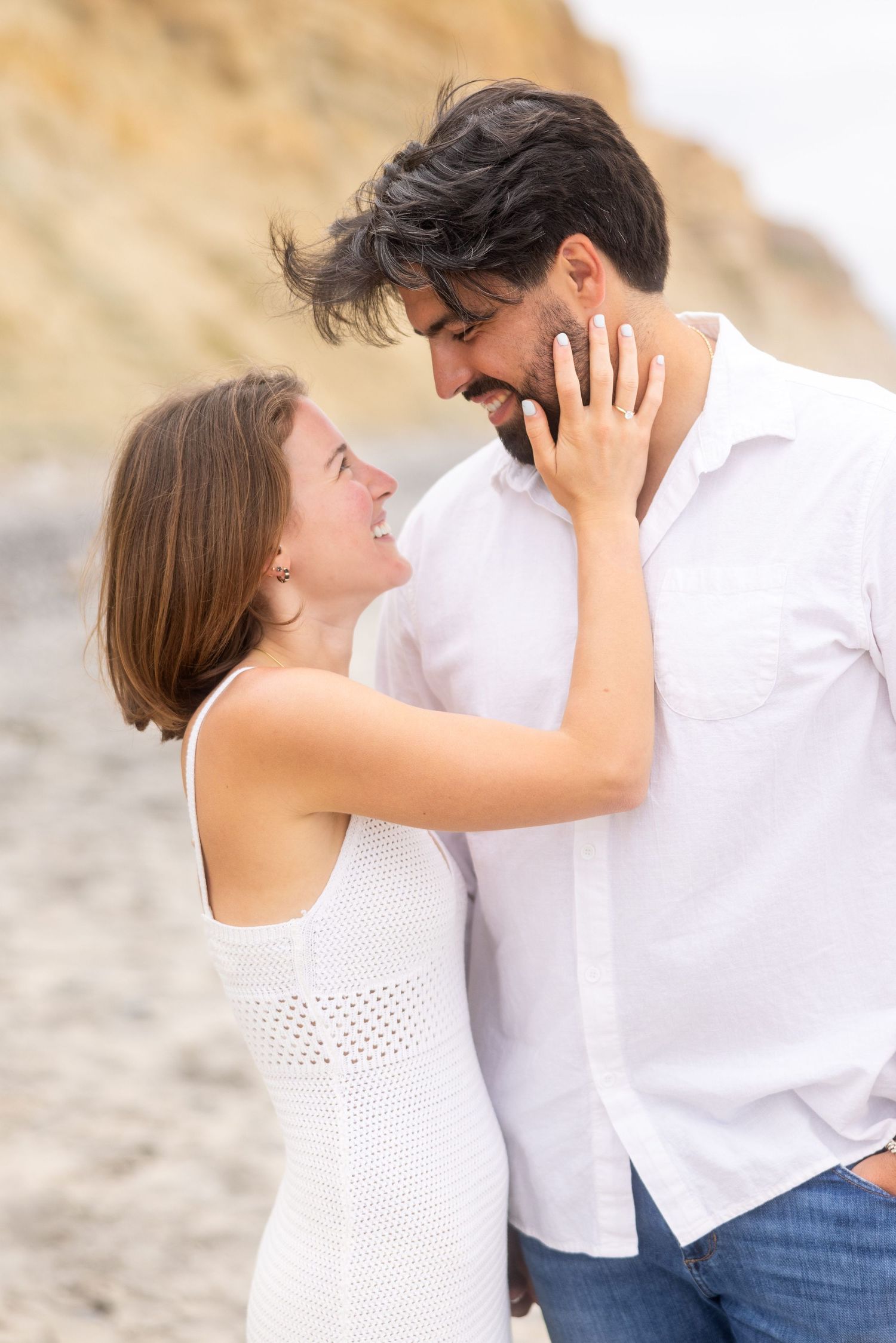 A romantic couple wearing white and blue embrace on a sandy beach during a sunny day.