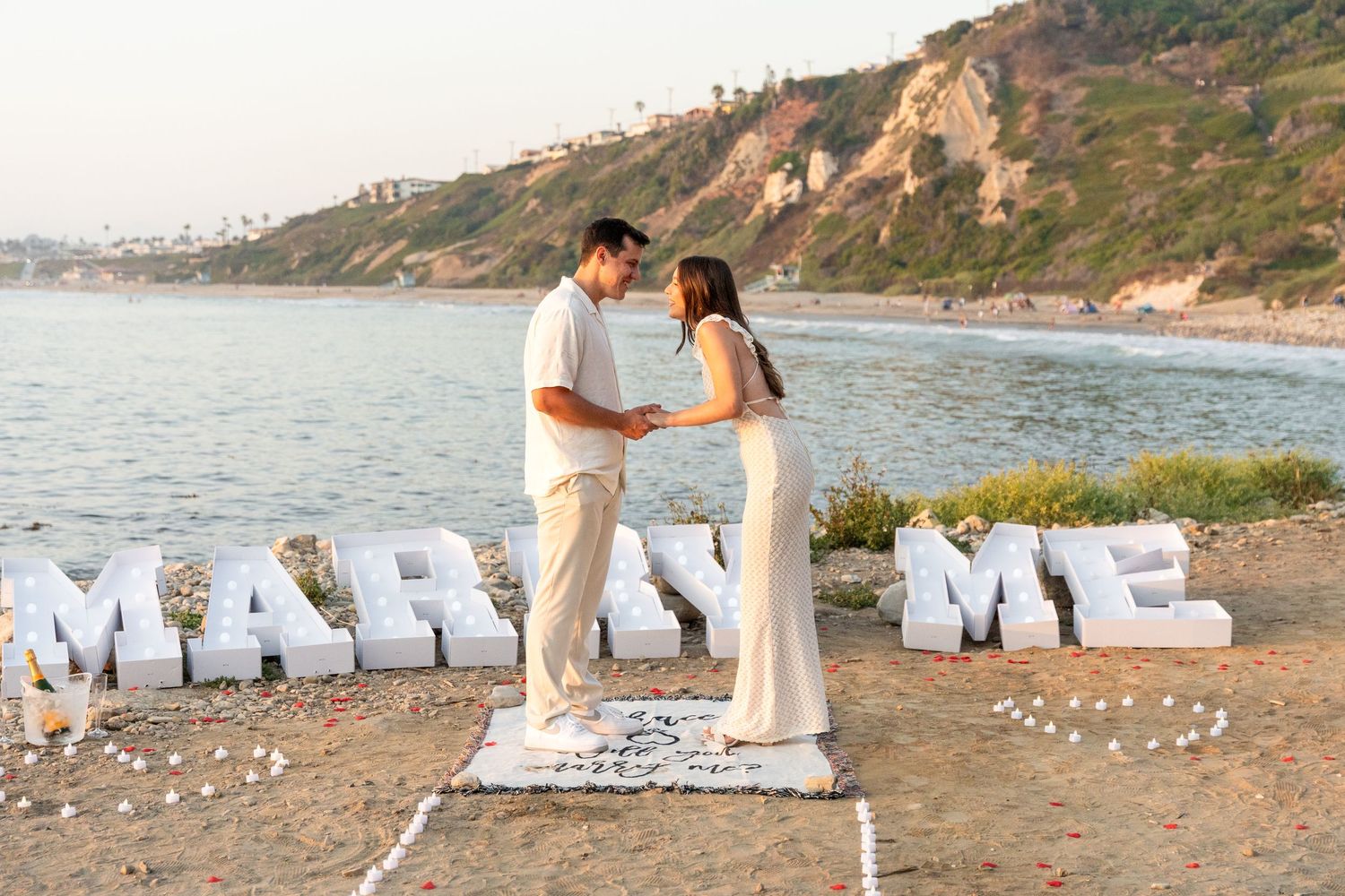 Couple stands near large white letter sign spelling out 'MARRY ME' on a scenic beach cliff during sunset.