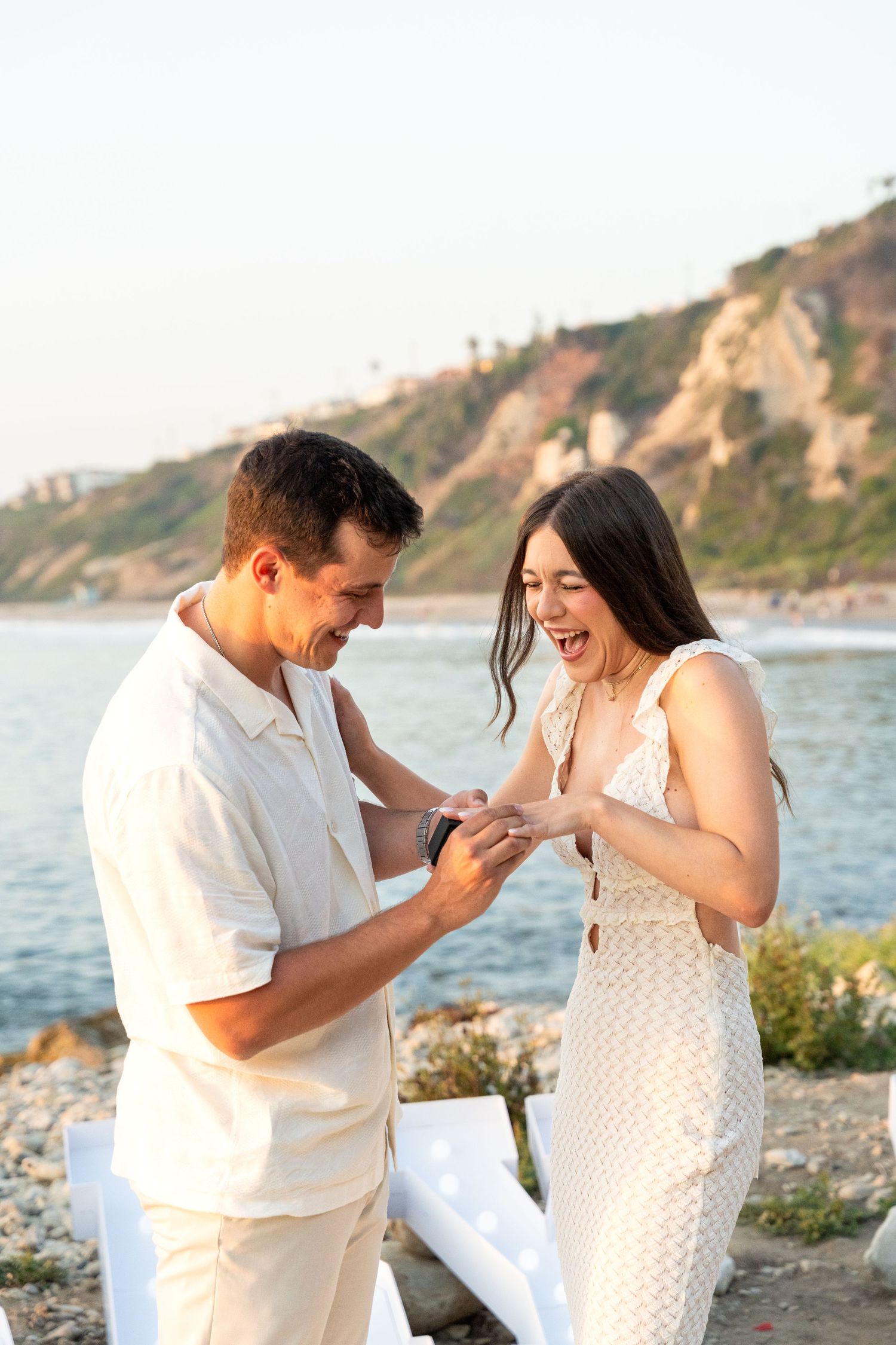 A couple shares a sweet moment during proposal on a scenic rocky beach at sunset.