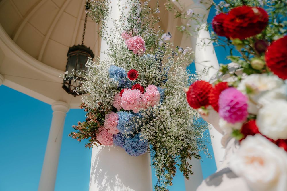 Wedding floral arrangements in pink and red hang from white gazebo ceiling against bright blue sky.