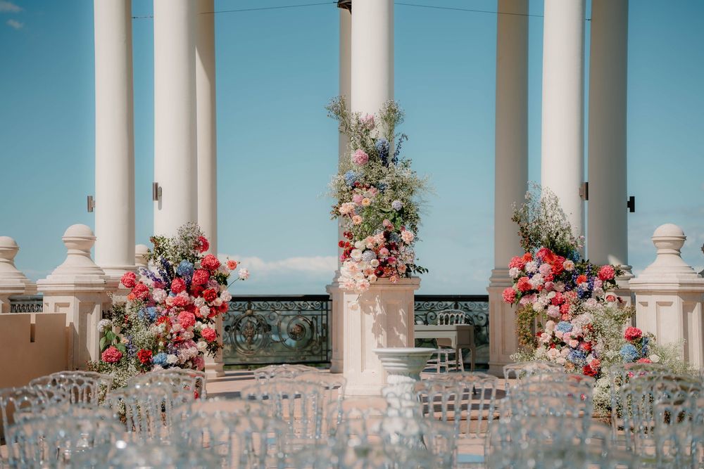 Elegant wedding ceremony setup with white columns and vibrant coral and red floral arrangements under a sunny blue sky.
