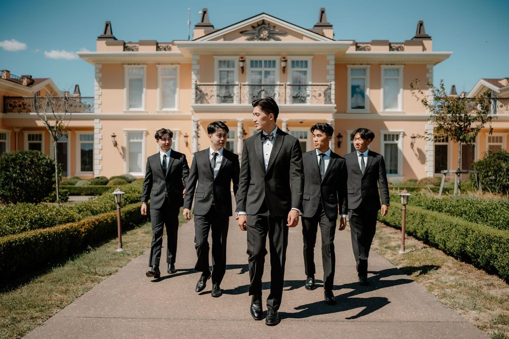 A row of groomsmen in matching gray suits walking along a grand mansion exterior on a sunny day.