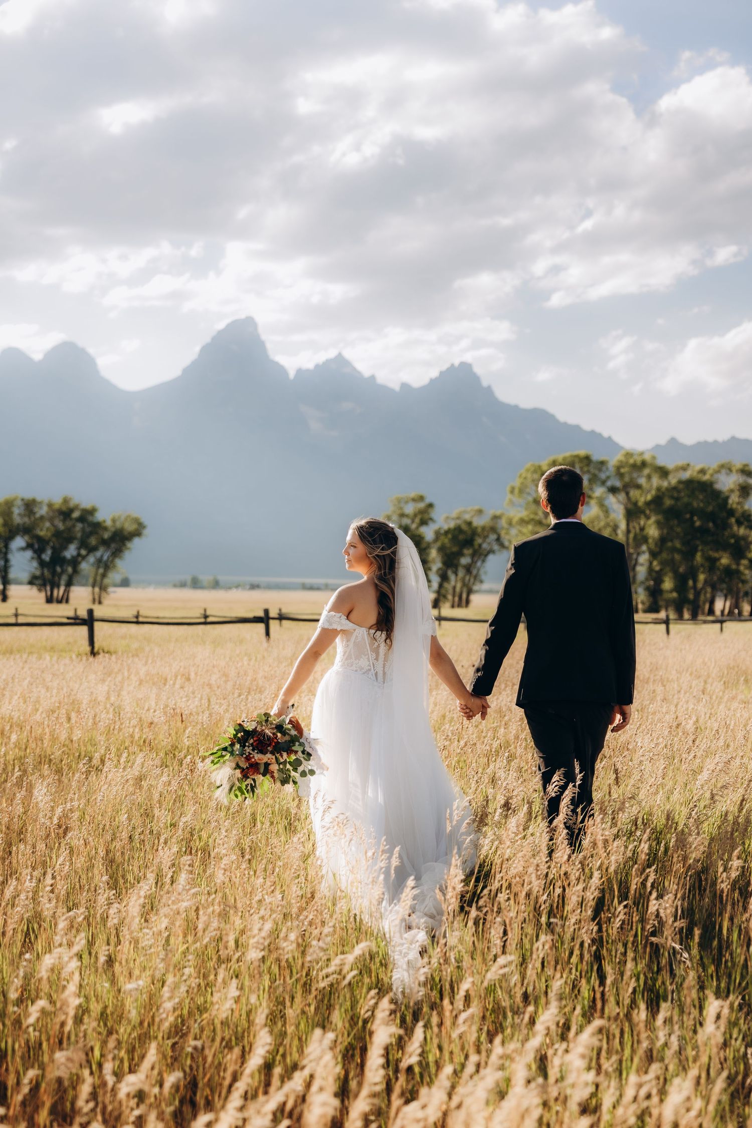 Newlywed couple walks hand in hand through a golden wheat field with misty mountains in the background.