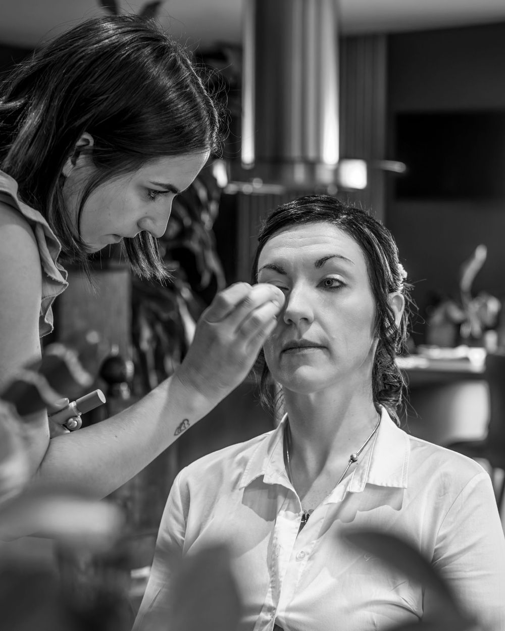 Black and white photo of a makeup artist applying makeup to a woman seated in a salon chair.