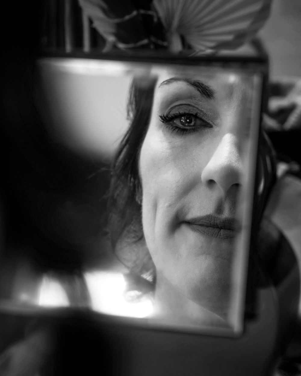 Black and white photo of a woman smiling, reflected in a small mirror with dramatic eye makeup.
