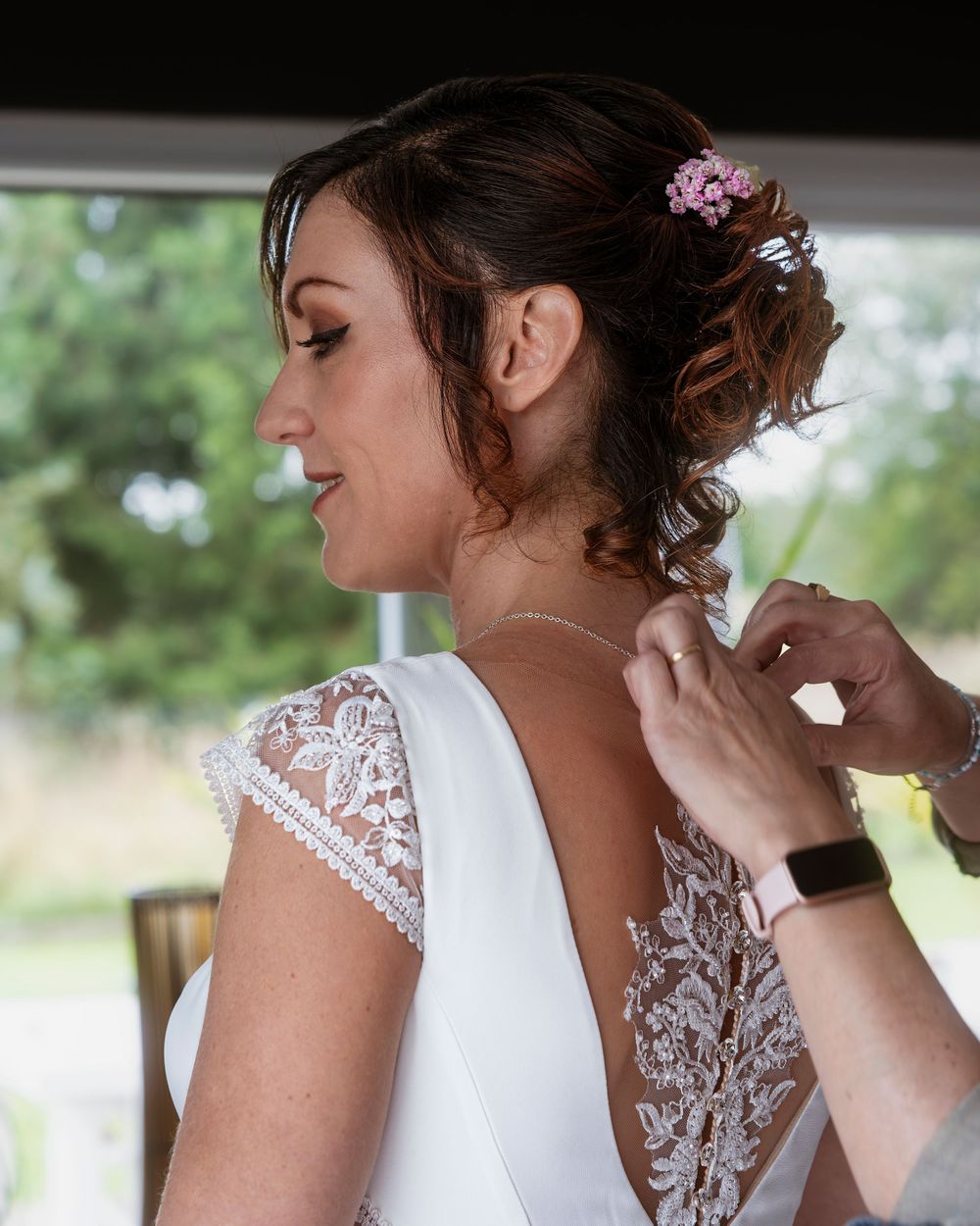 Bride in lace-back wedding dress having necklace fastened, with pink flowers in her updo hairstyle.