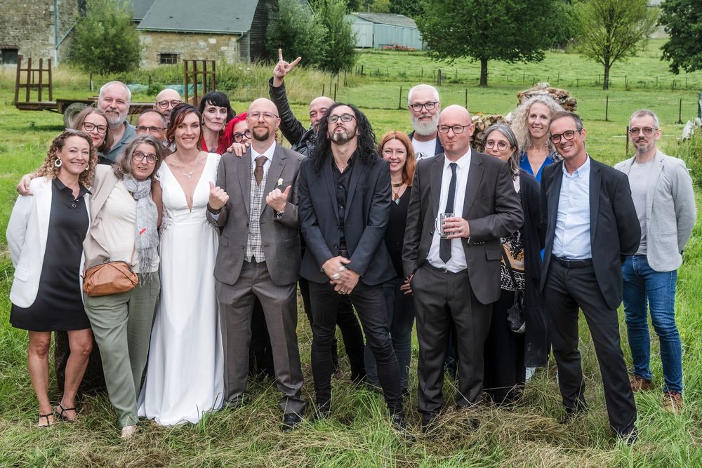 A group of people dressed in formal attire pose together outdoors at a wedding celebration in a countryside setting.