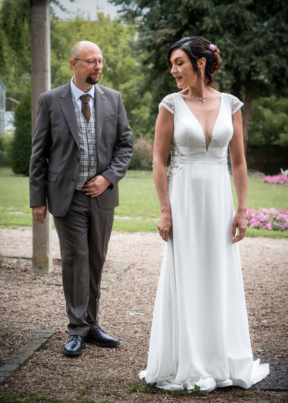 A romantic wedding couple holds hands on a gravel path surrounded by lush green gardens and flowers.