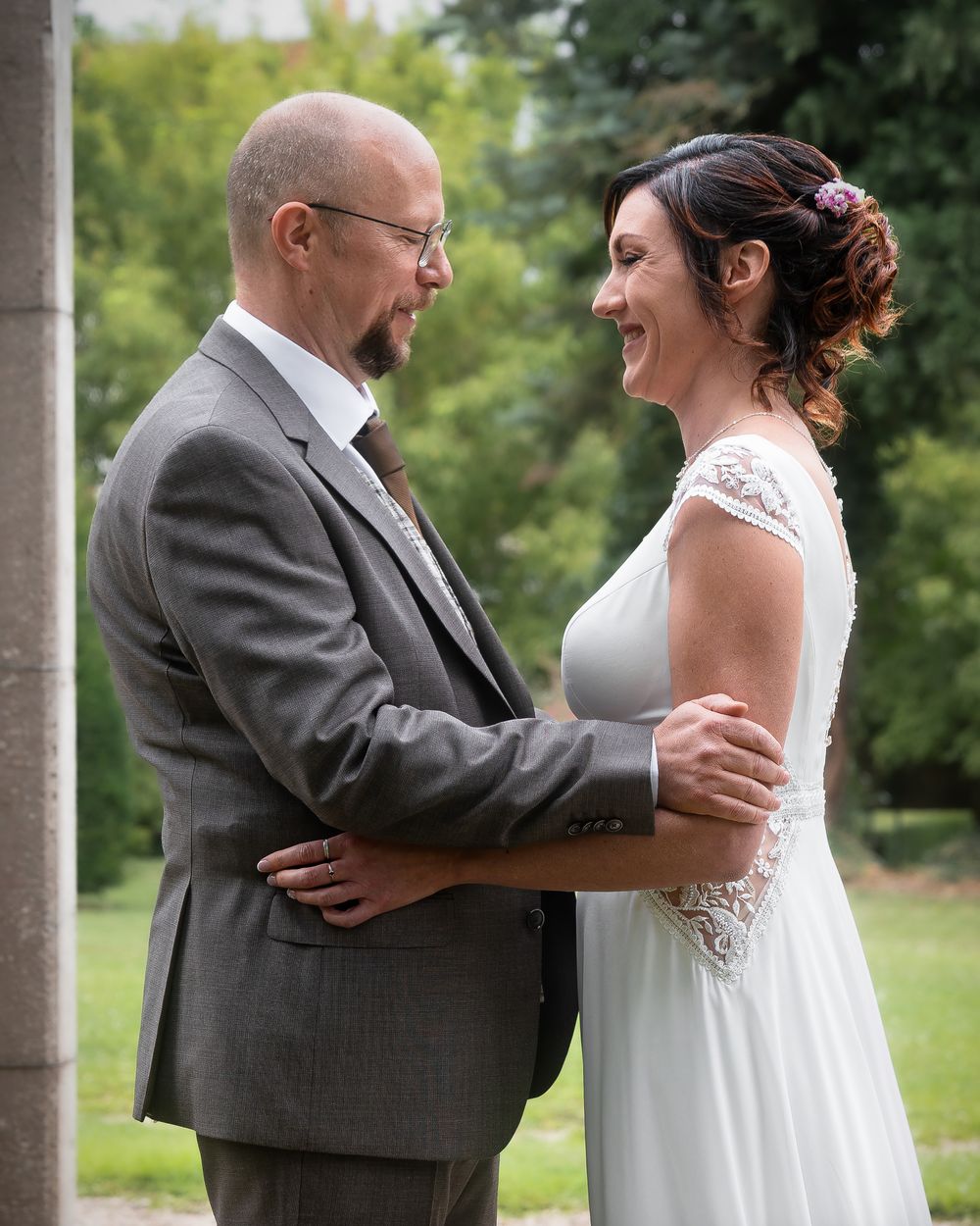 Bride in white lace dress and groom in gray suit share joyful laughter during their wedding portraits outdoors.