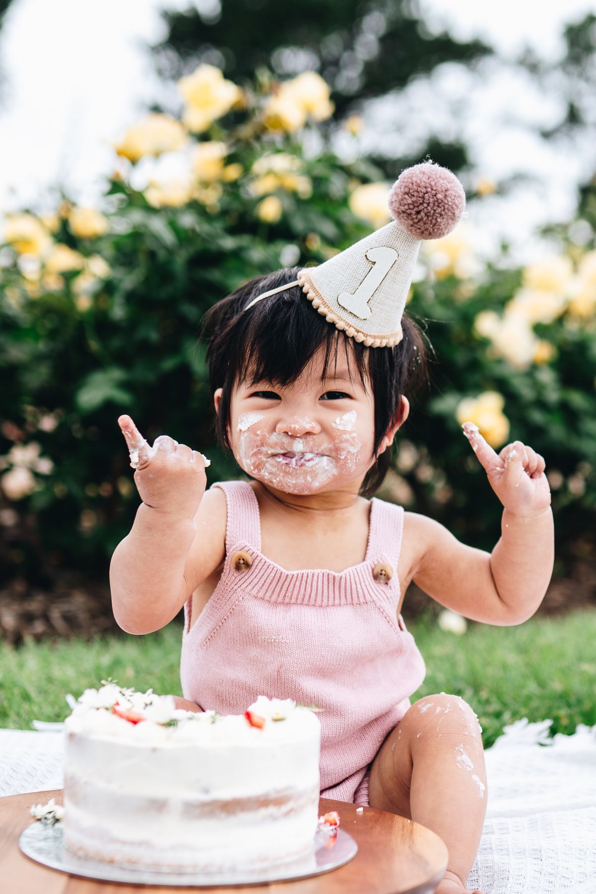 Cake smash photo session featuring a white cake on black pedestal in sunny outdoor Melbourne park celebrating first birthday.