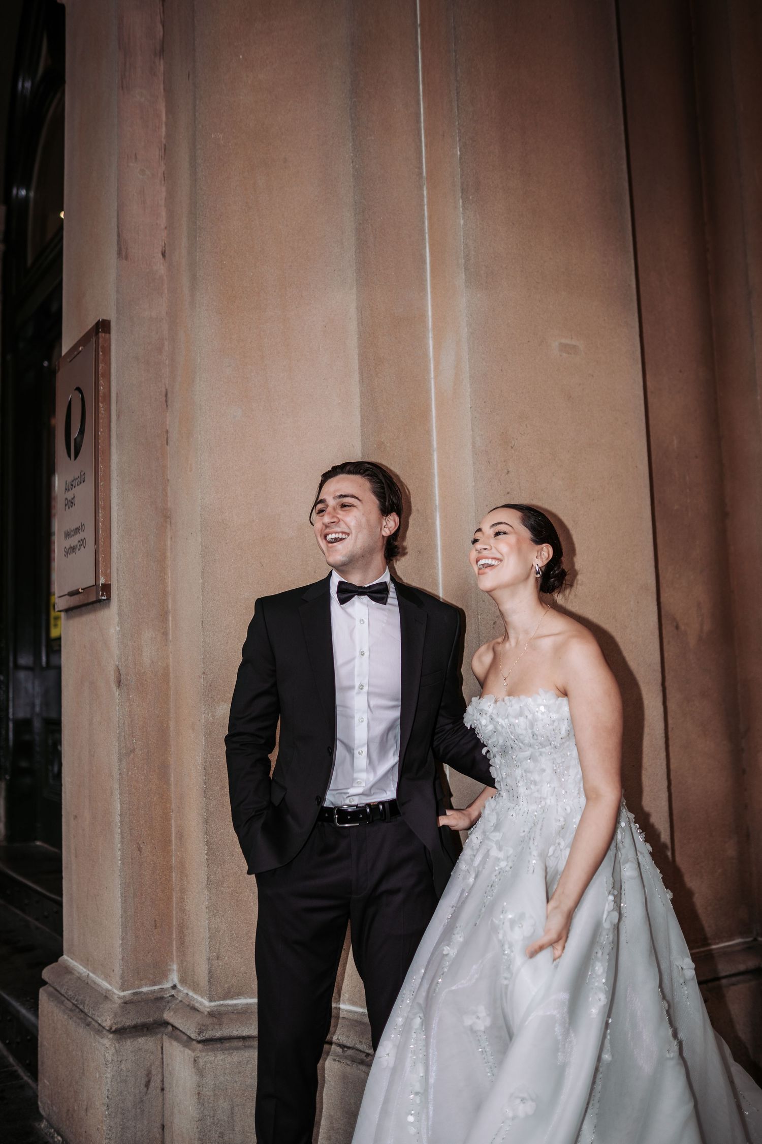 A bride and groom share a joyful laugh while standing against a classic stone pillar at their wedding.