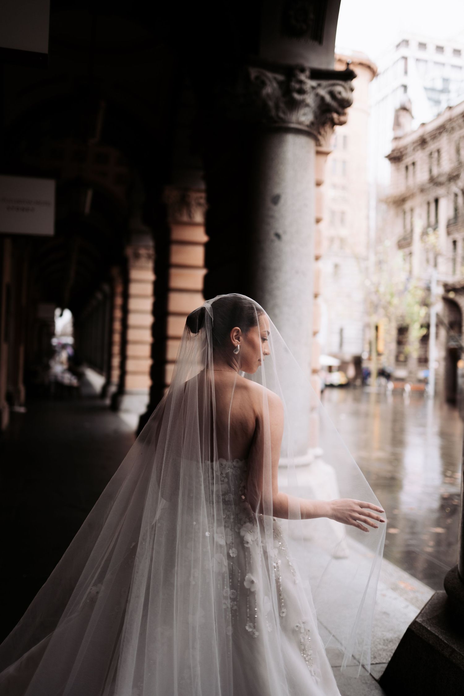 A bride in a flowing white wedding dress and veil stands beneath stone arches in an elegant corridor.