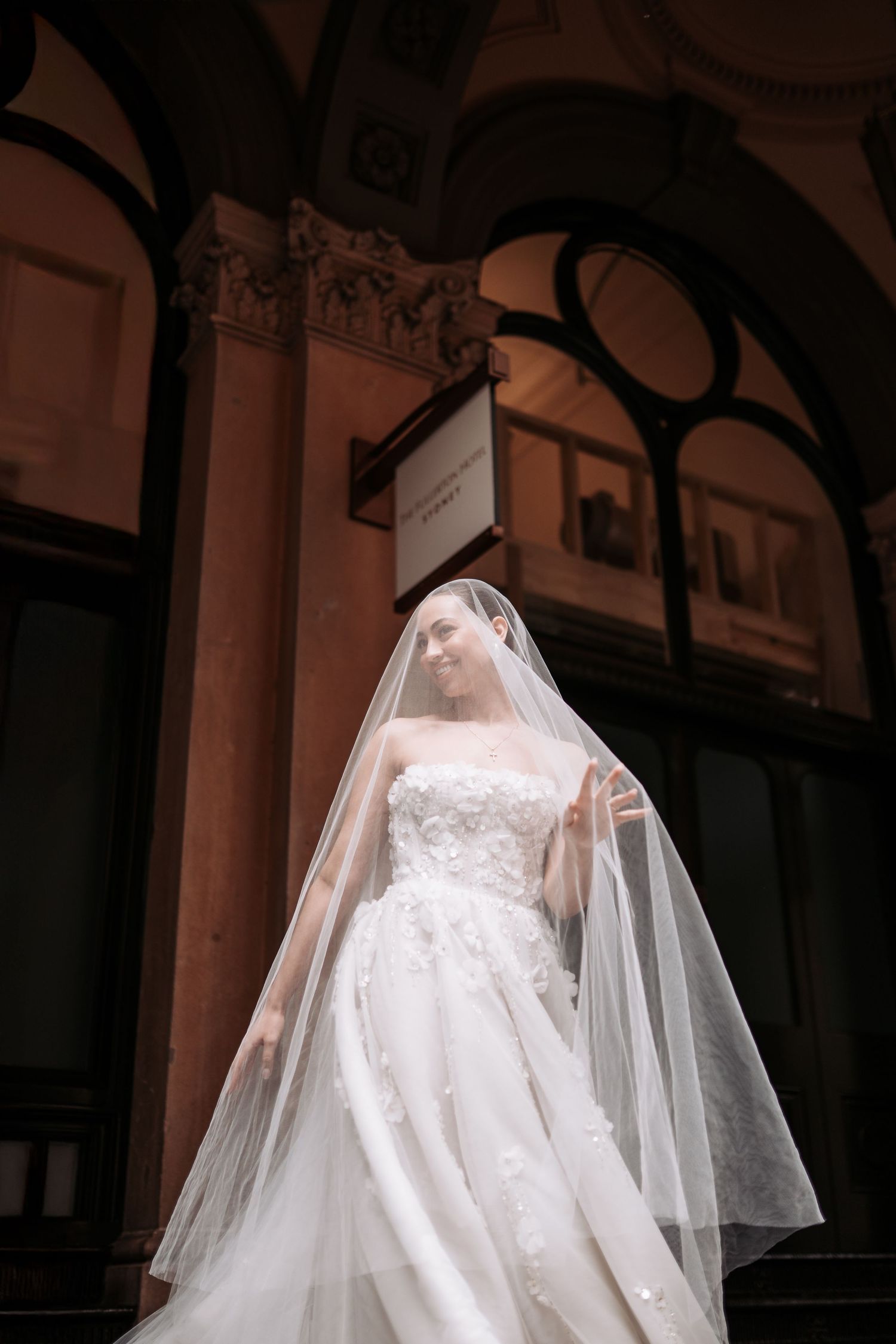 A bride in a white lace wedding dress and flowing veil stands in an elegant arched hallway.