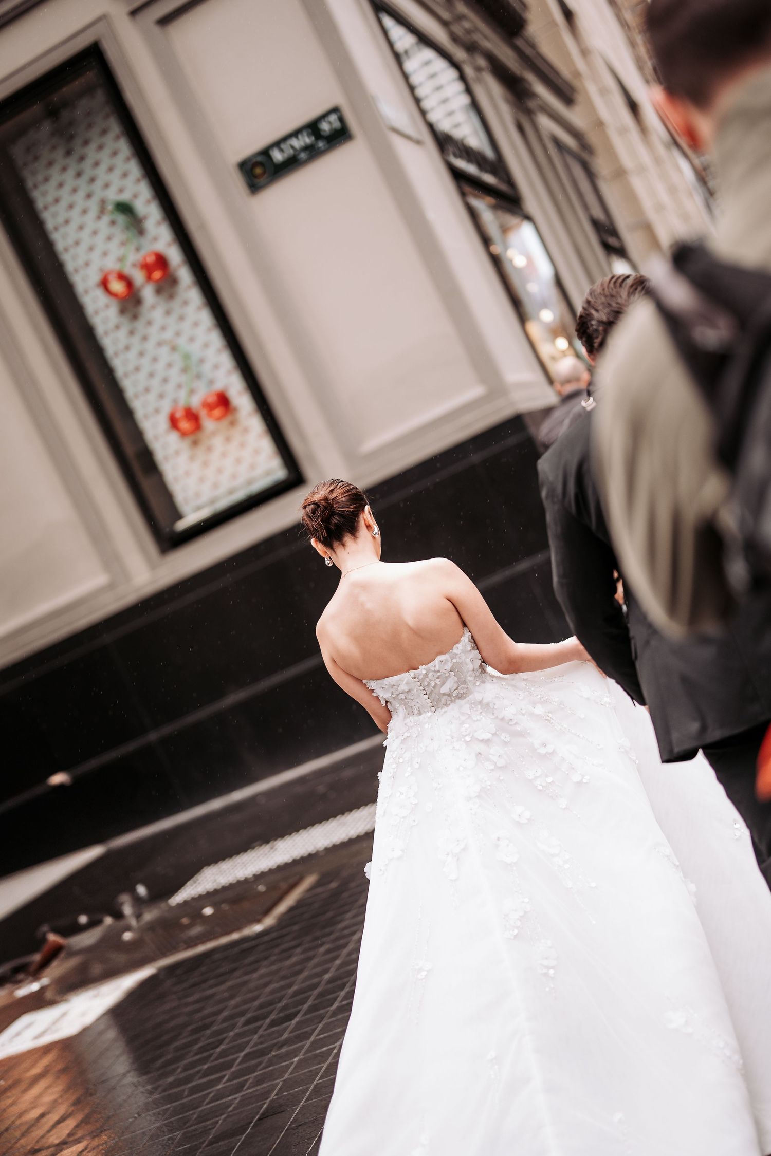 A bride in a strapless white wedding gown stands outside a building with textured glass doors.