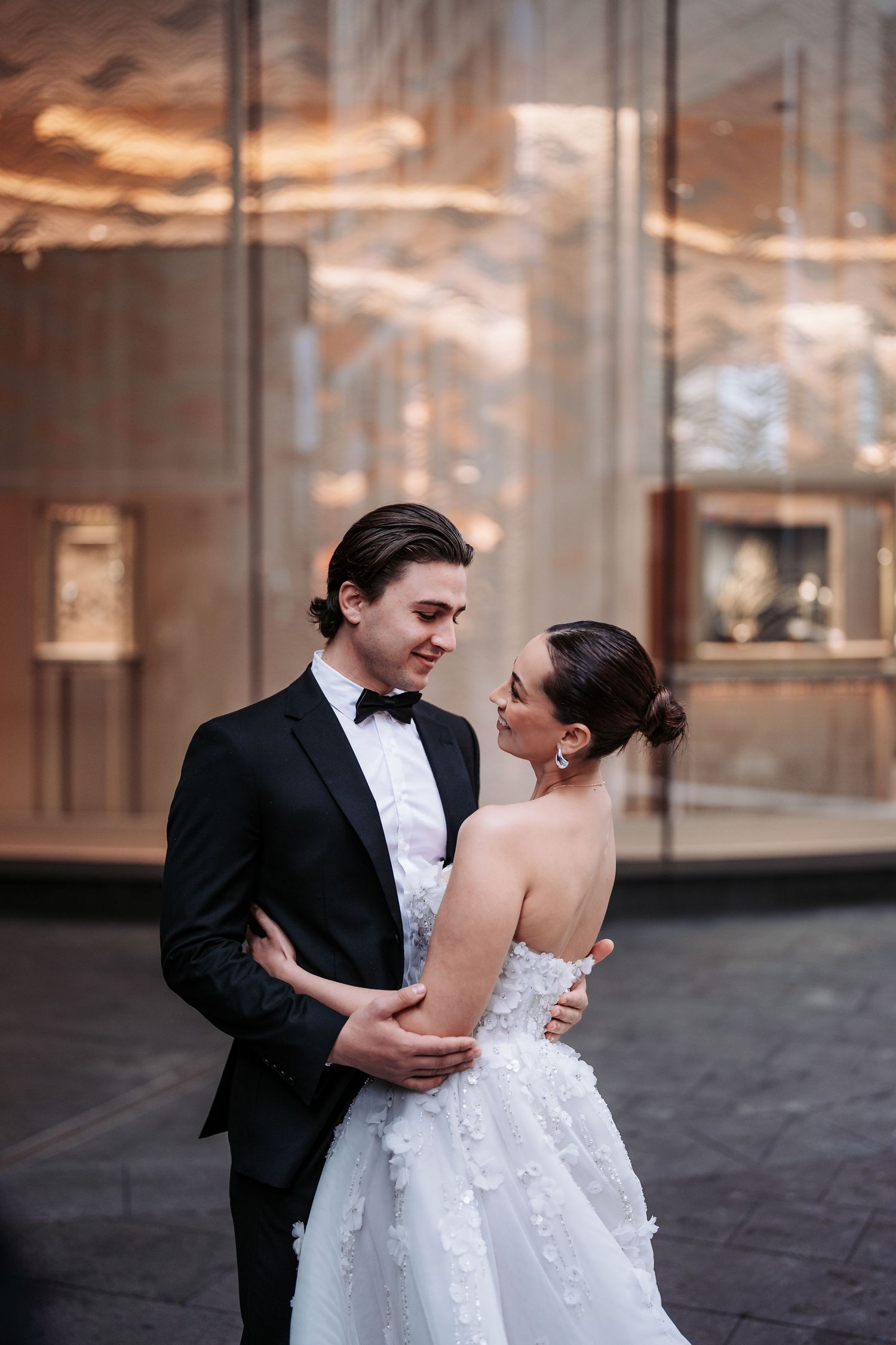 A romantic wedding couple embraces in formal attire in front of an elegant glass display.