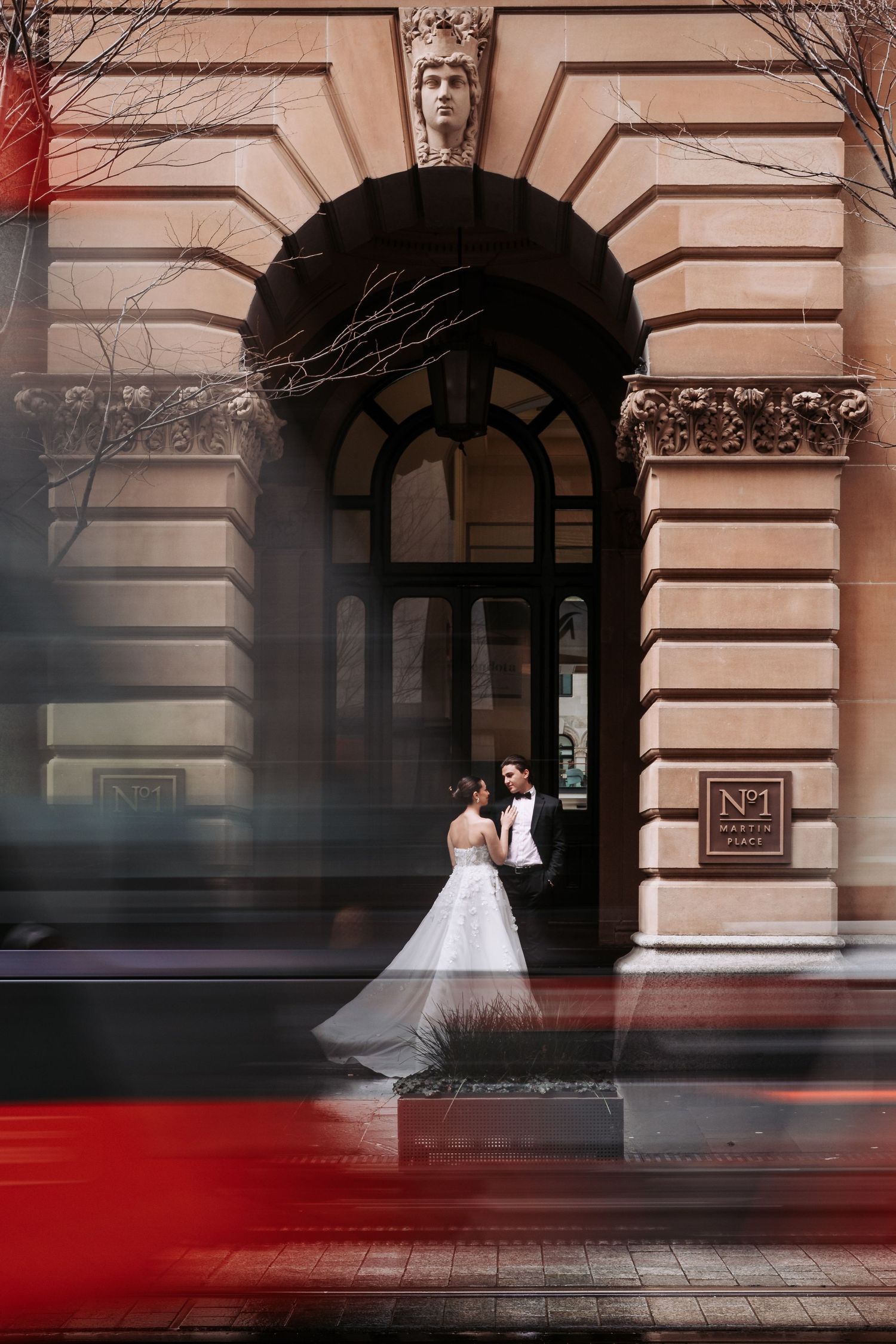 Elegant wedding couple photographed against classic building facade with dramatic red reflection on sidewalk.