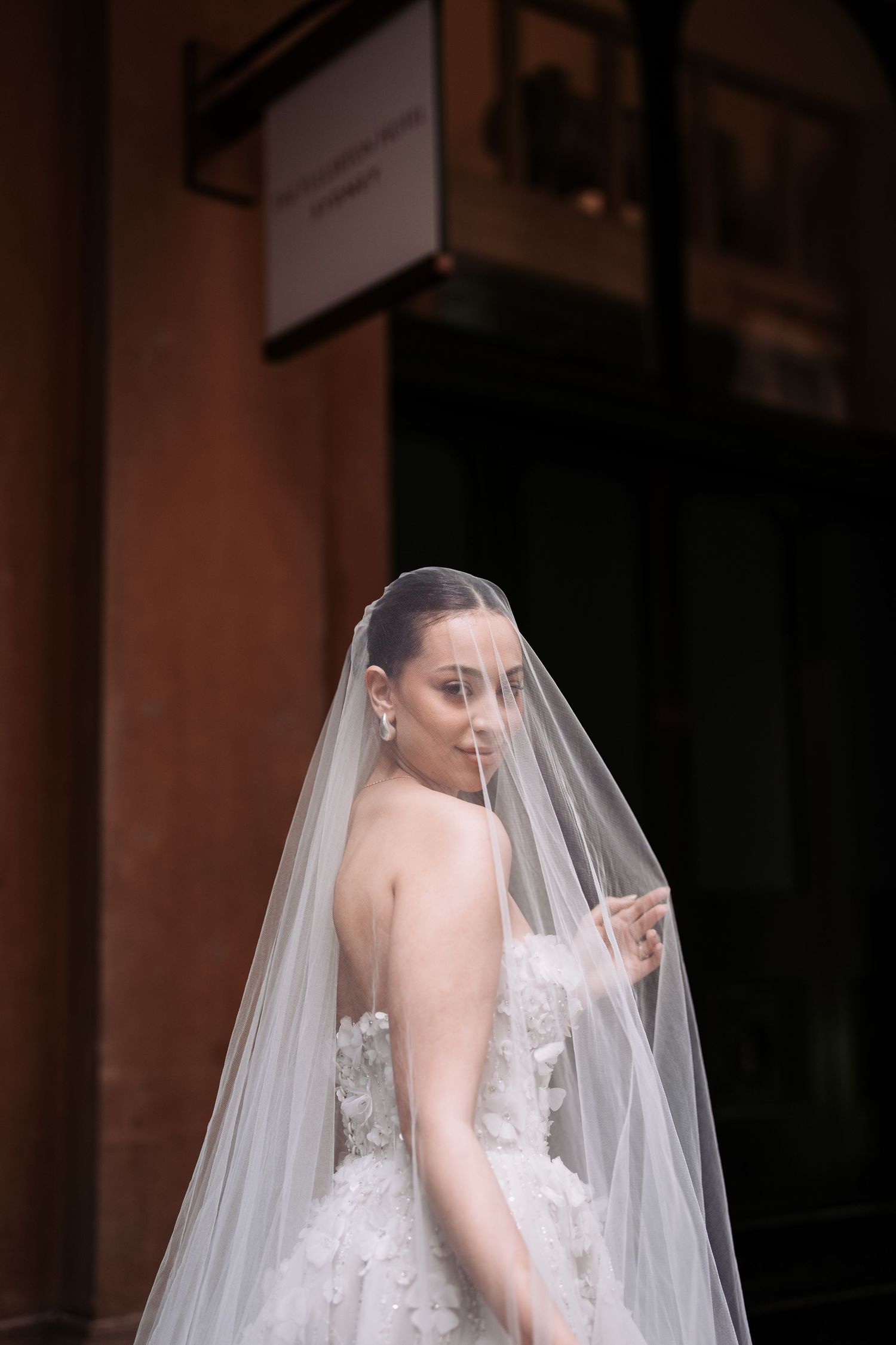 A bride in a strapless lace wedding gown looks over her shoulder through a flowing long veil during a photoshoot.