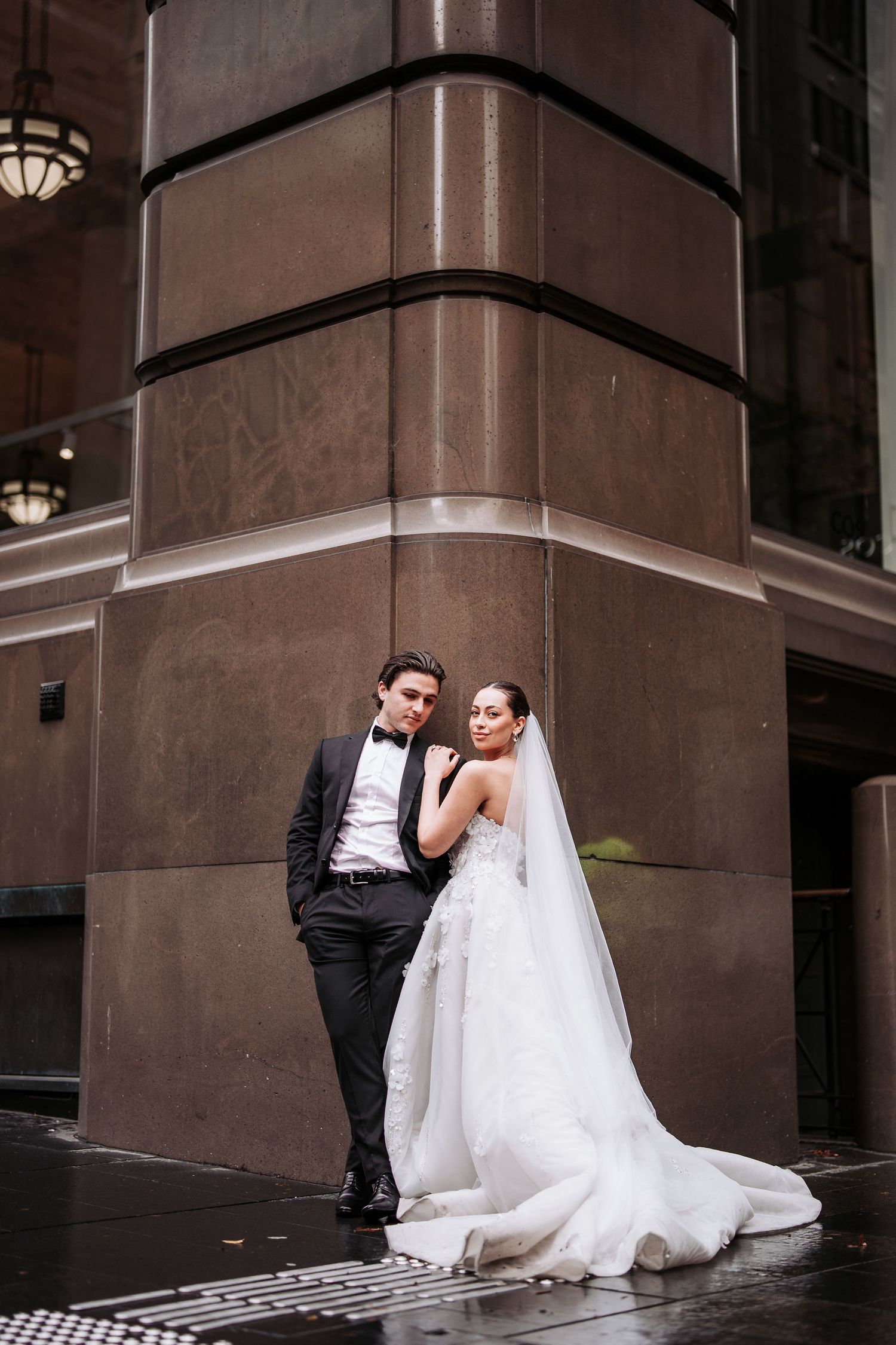 A couple in wedding attire pose elegantly against a brown stone building corner in an urban setting.