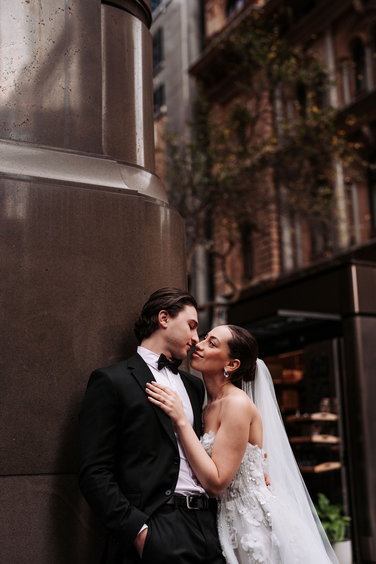 A romantic couple in formal wedding attire embraces against a stone building wall.