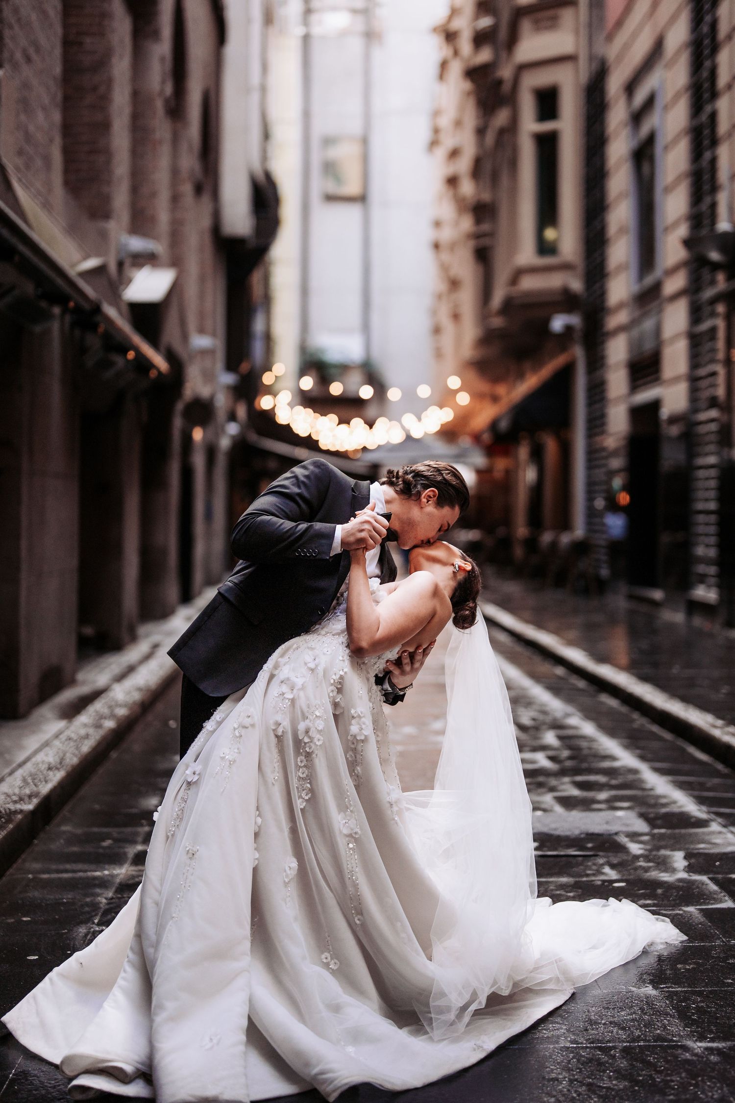 A romantic wedding couple shares intimate moments on a cobblestone street while laying on the ground in an urban setting.