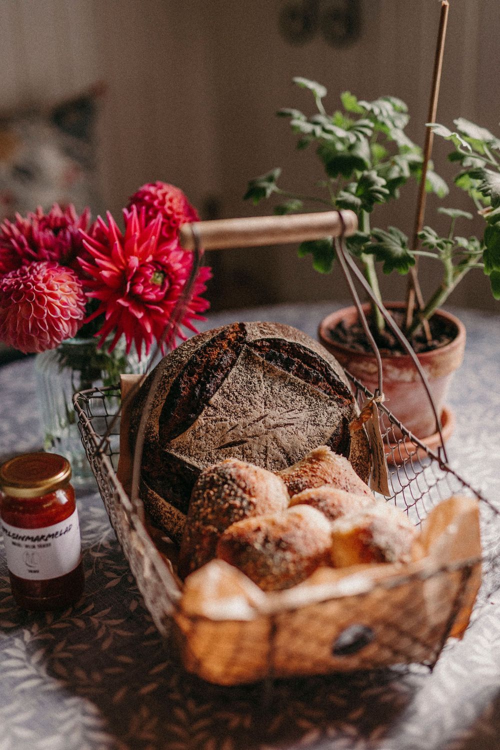 Rustic bread basket with fresh rolls and dark bread alongside red carnations and a small potted plant on a textured tablecloth.