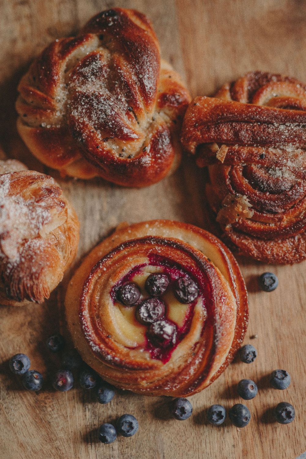 Fresh baked sweet pastry rolls with blueberries scattered around on a rustic wooden surface dusted with powdered sugar.