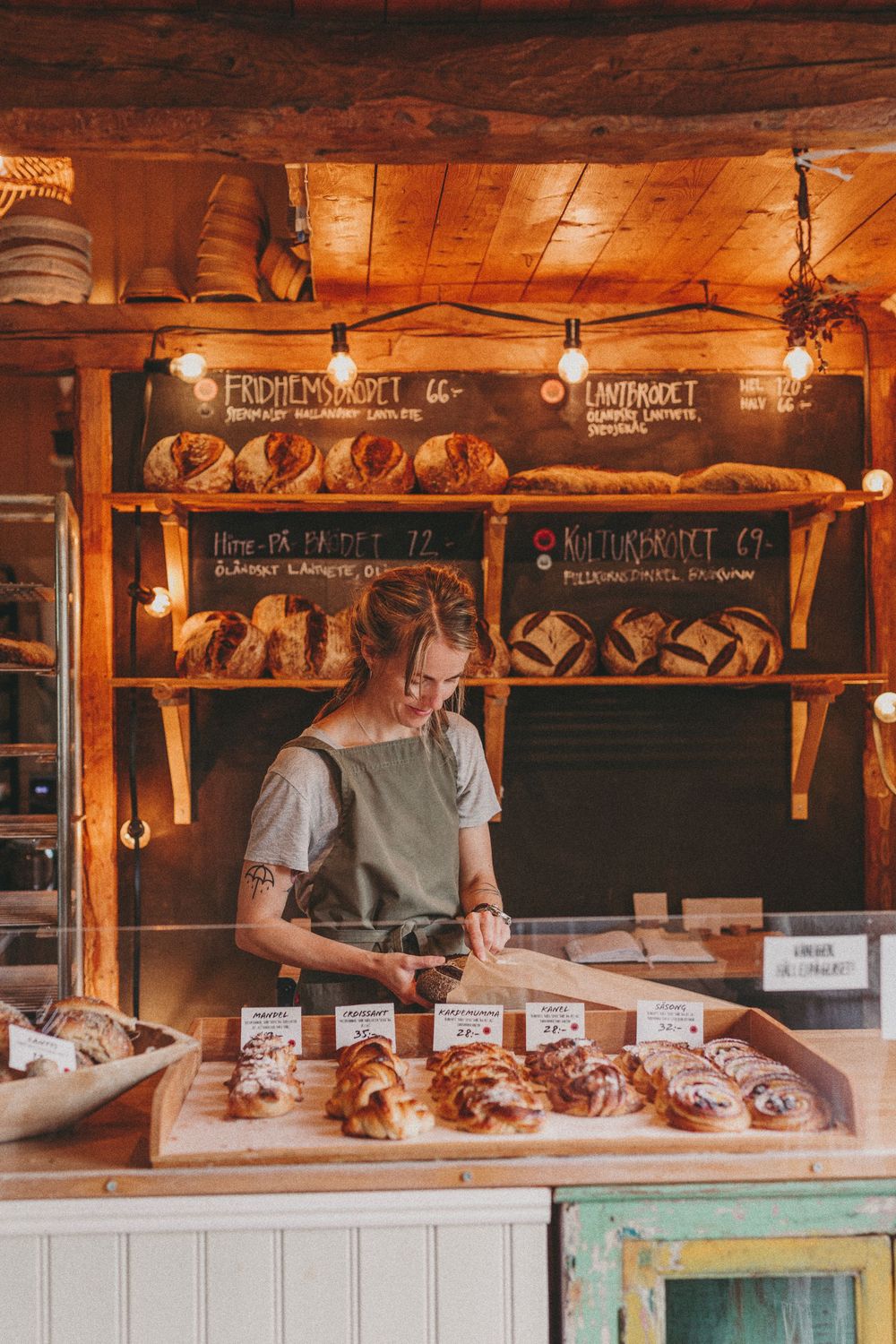 Baker arranges fresh pastries and bread on wooden shelves in warm rustic bakery with chalkboard menus.