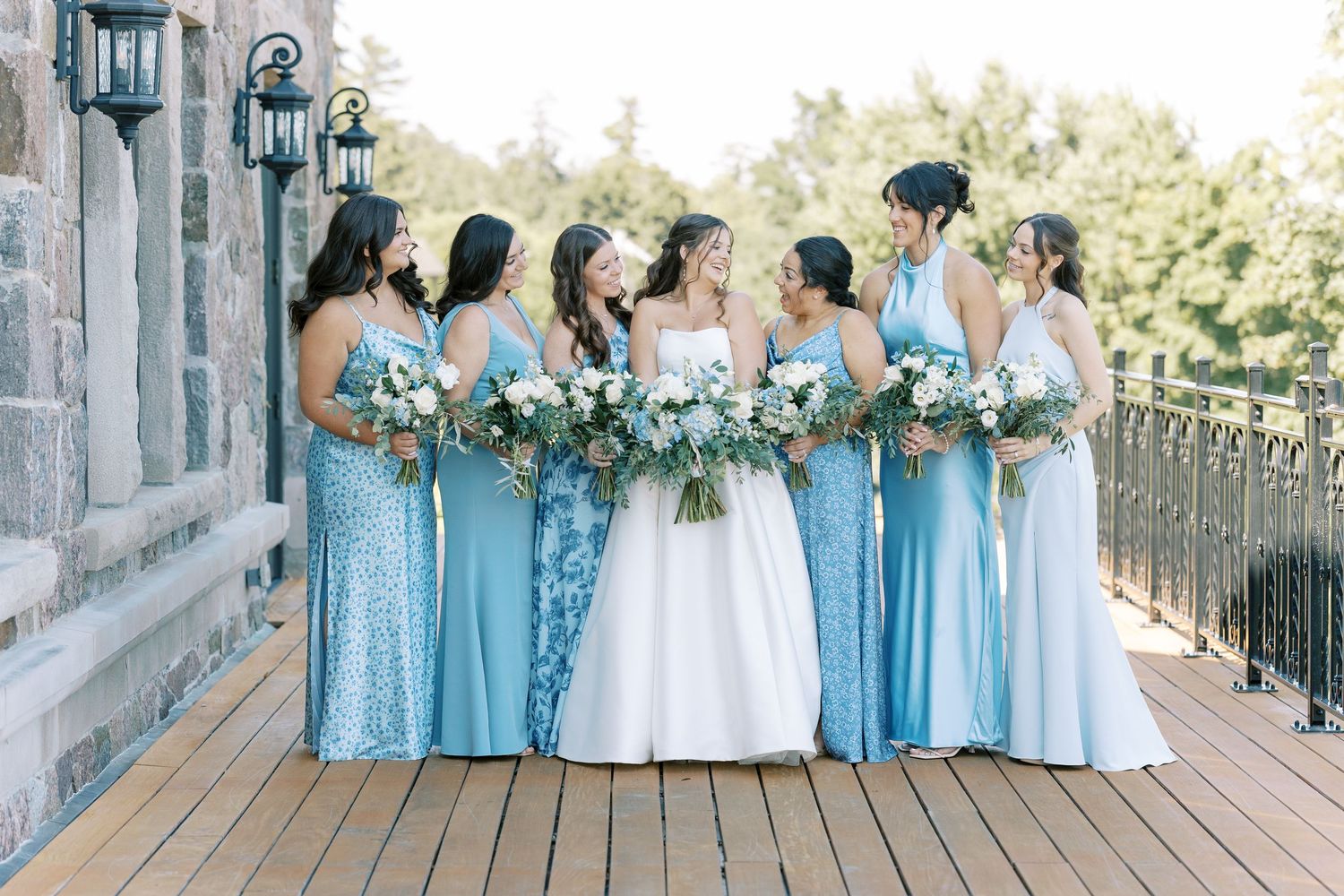 Wedding party wearing light blue bridesmaid dresses poses together on outdoor balcony with stone columns.