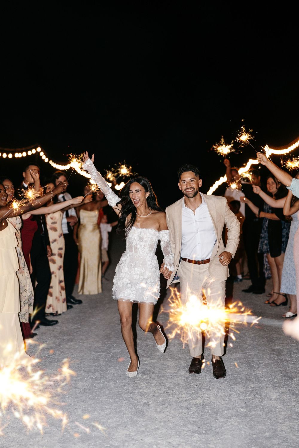 Group of celebrants dance and hold sparklers on a beach during a nighttime wedding party celebration.
