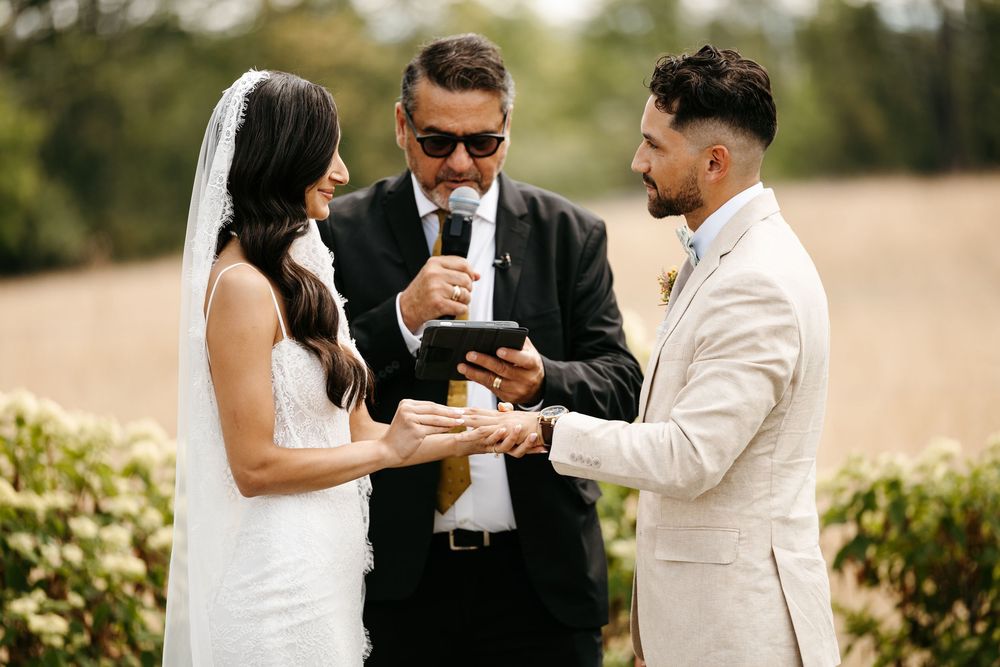 An outdoor wedding ceremony takes place in a garden with the officiant and couple sharing a lighthearted moment.