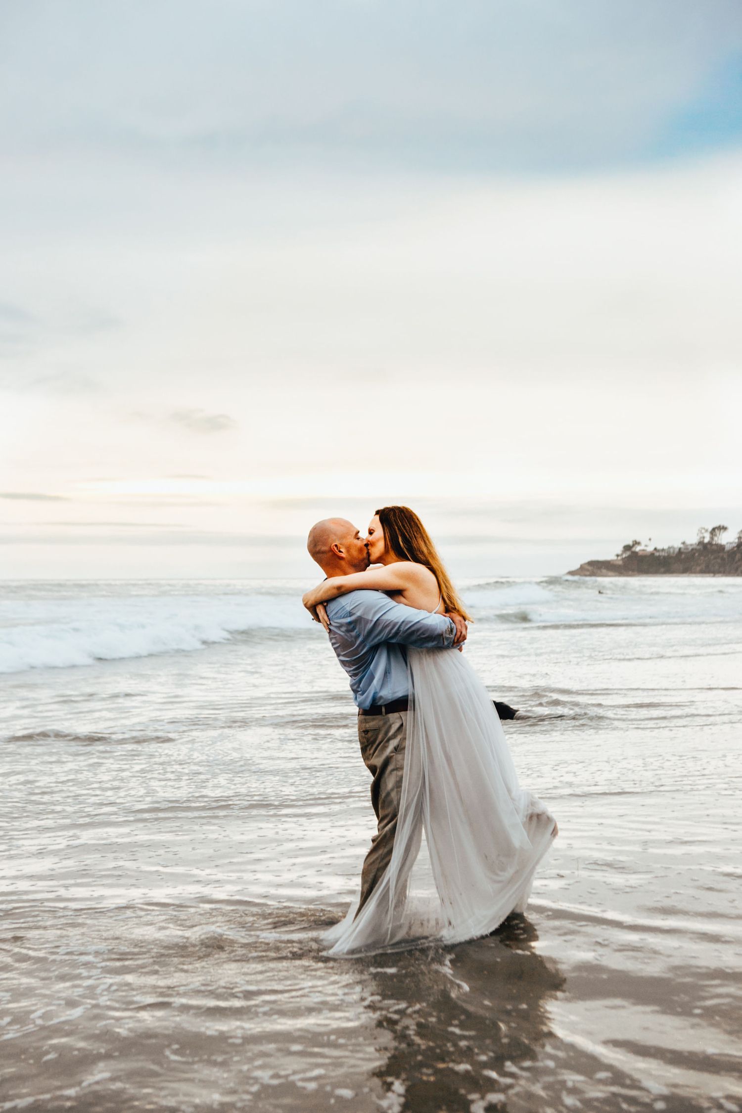 Romantic couple dancing in flowing white dress along the ocean shore at sunset in ethereal beach photoshoot in Laguna Beach, California.