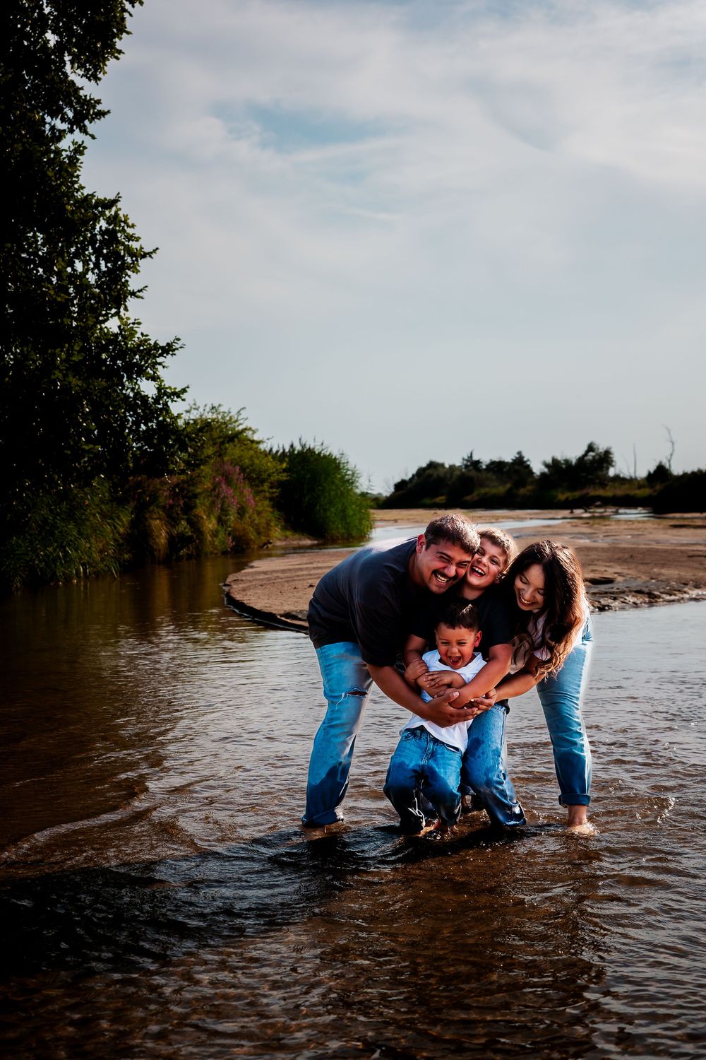 Family together while standing in shallow water at the edge of a river on a cloudy day during a Kearney, Nebraska family photography session