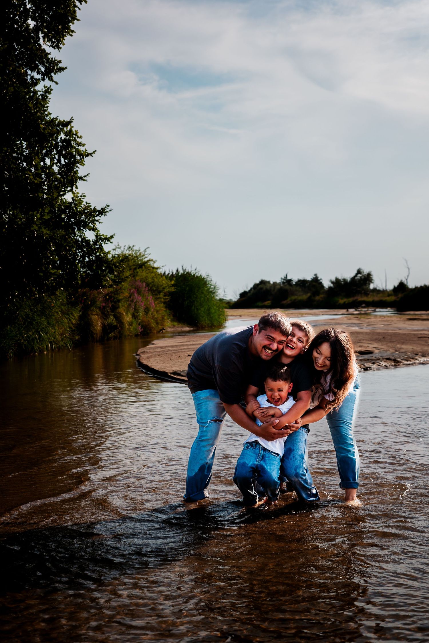 Family together while standing in shallow water at the edge of a river on a cloudy day during a Kearney, Nebraska family photography session