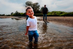 A joyful child plays in shallow water at the Platte River in Central Nebraska while an adult figure stands in the background.