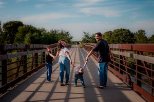 Family holding hands while walking together on a wooden bridge path outside Kearney, Nebraska, during golden hour with trees in background.