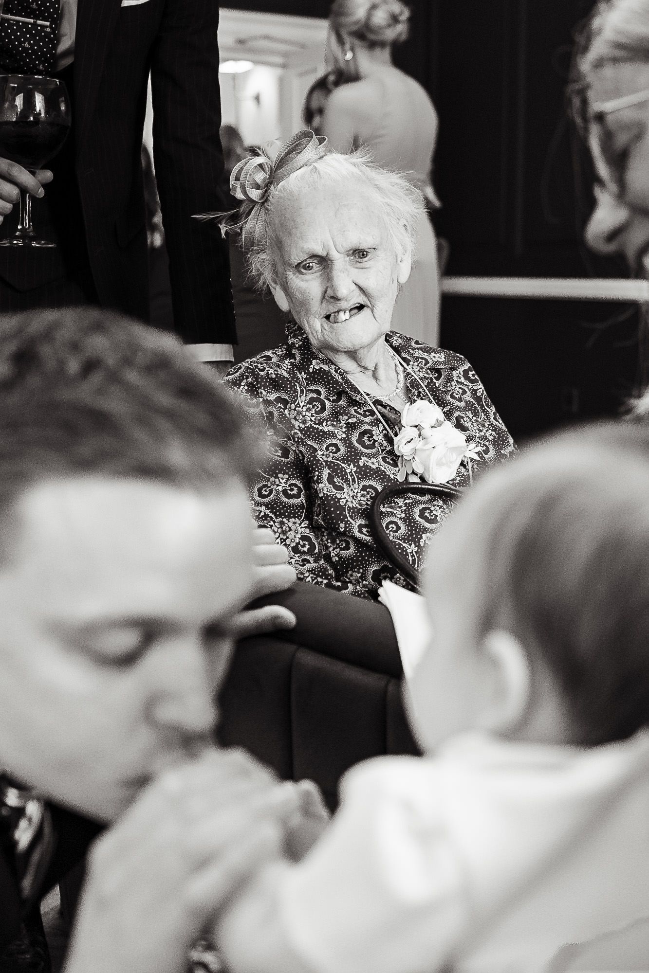 Elderly woman in patterned dress smiles at wedding celebration while guests mingle in black and white photograph.