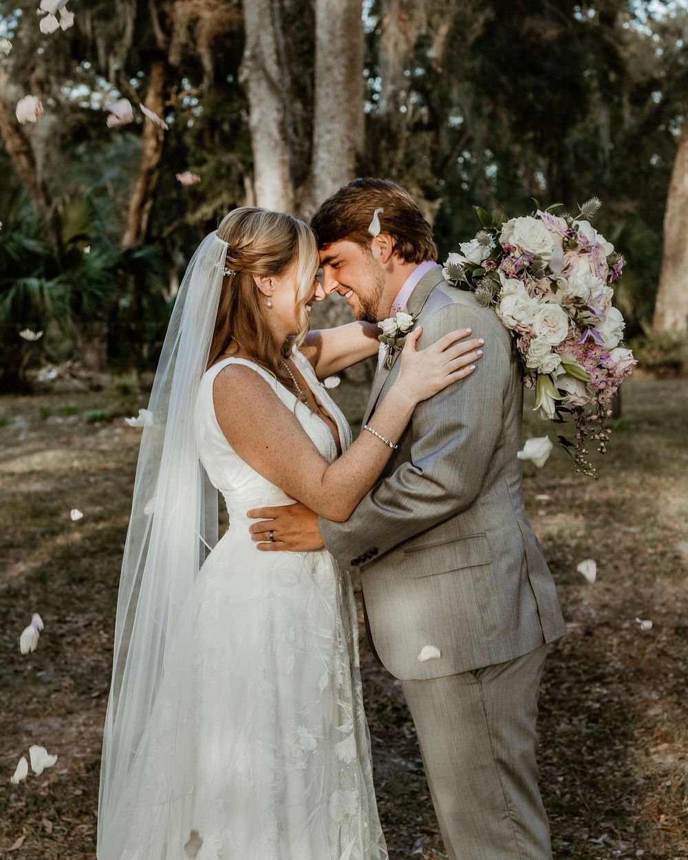 A tender moment between a couple in grey and white wedding attire surrounded by trees in an outdoor forest setting.